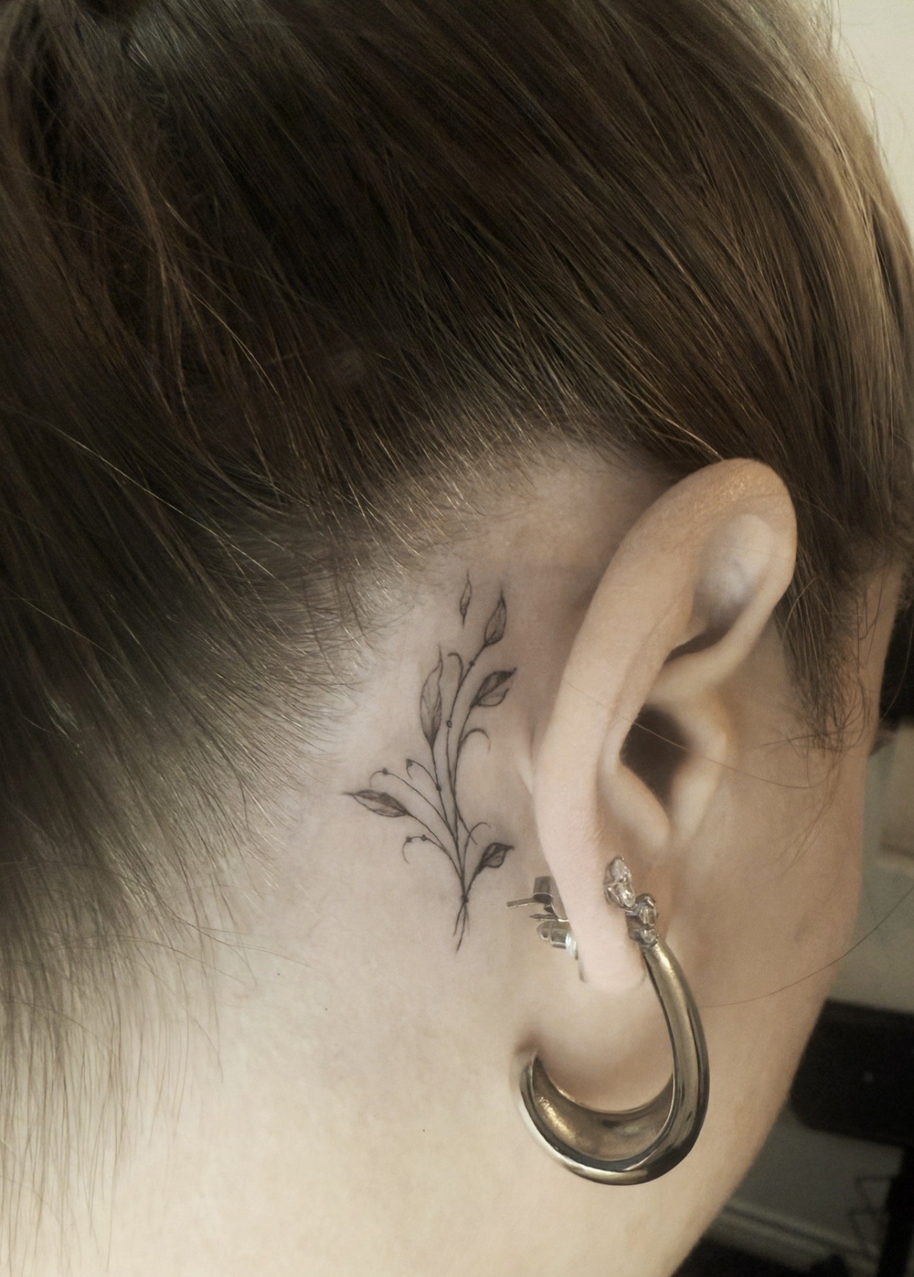 Close-up of a person's head showing brown short hair, a floral tattoo behind the ear, and multiple piercings including large hoop earrings and a pin with small charms.
