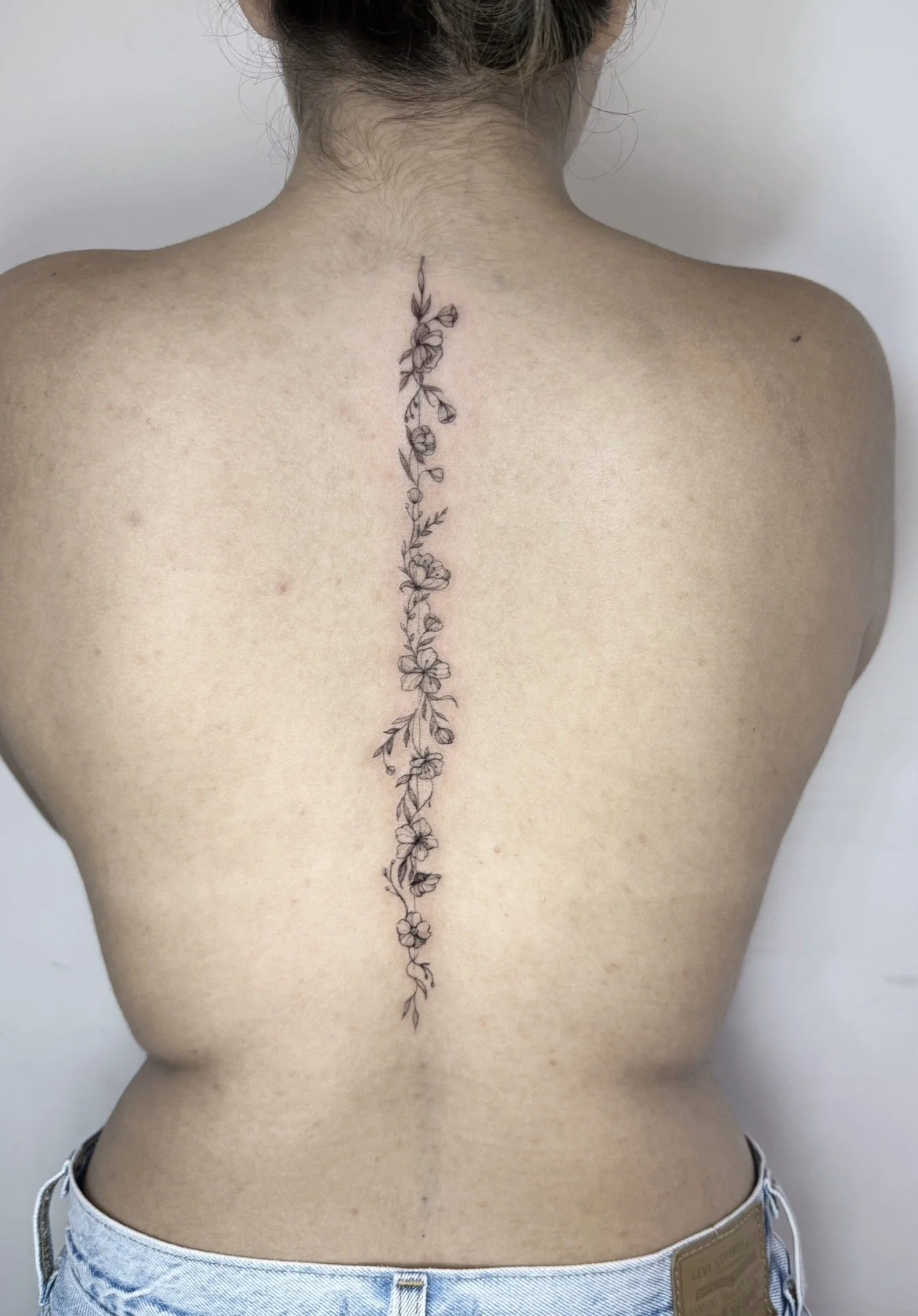 Back of a person with a floral tattoo running vertically along the spine, wearing light blue jeans.