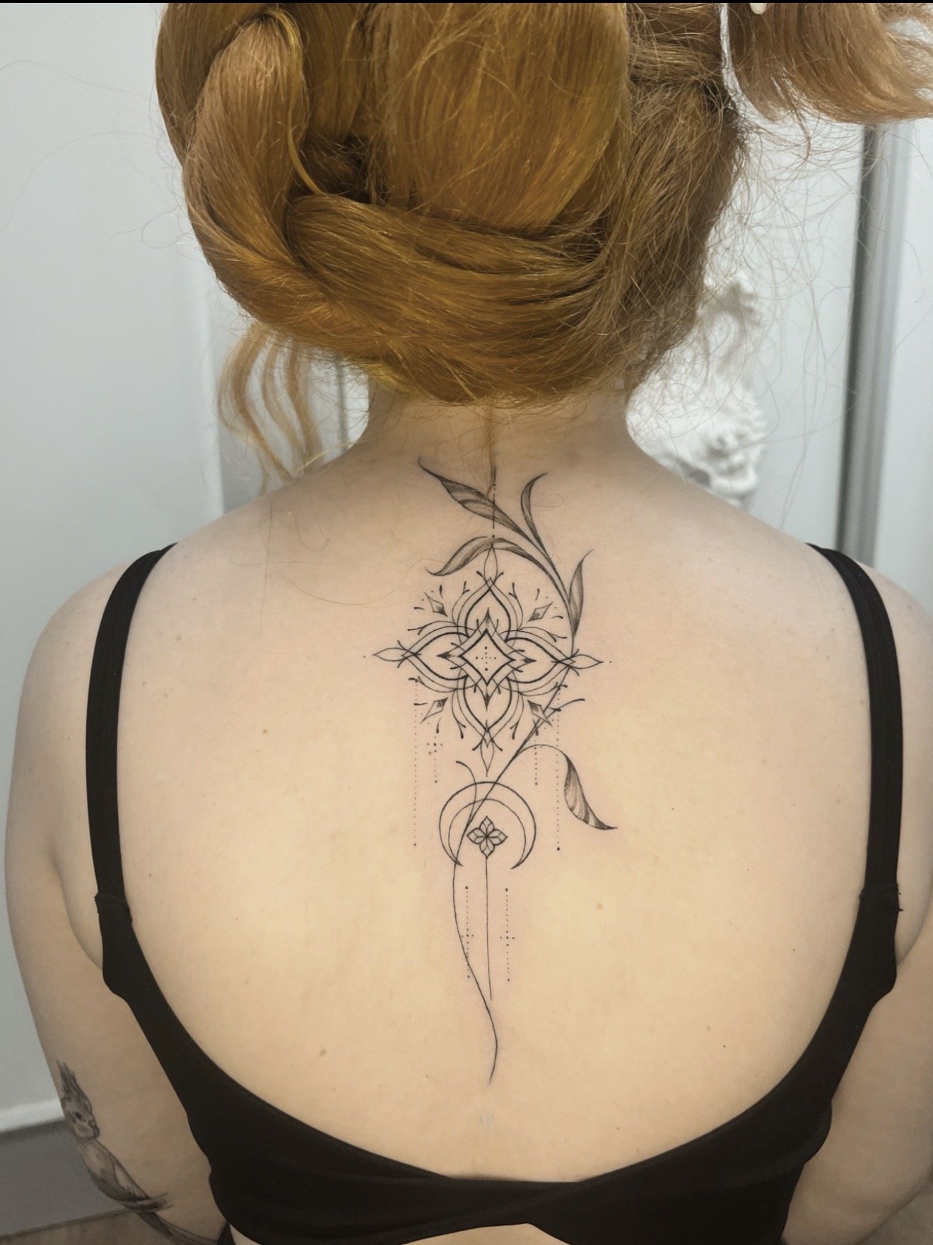 Back of a woman with red hair styled in a braid, wearing a black top with thin straps, showing a large, intricate, floral-themed tattoo on her upper back extending from her neck to her mid-back.