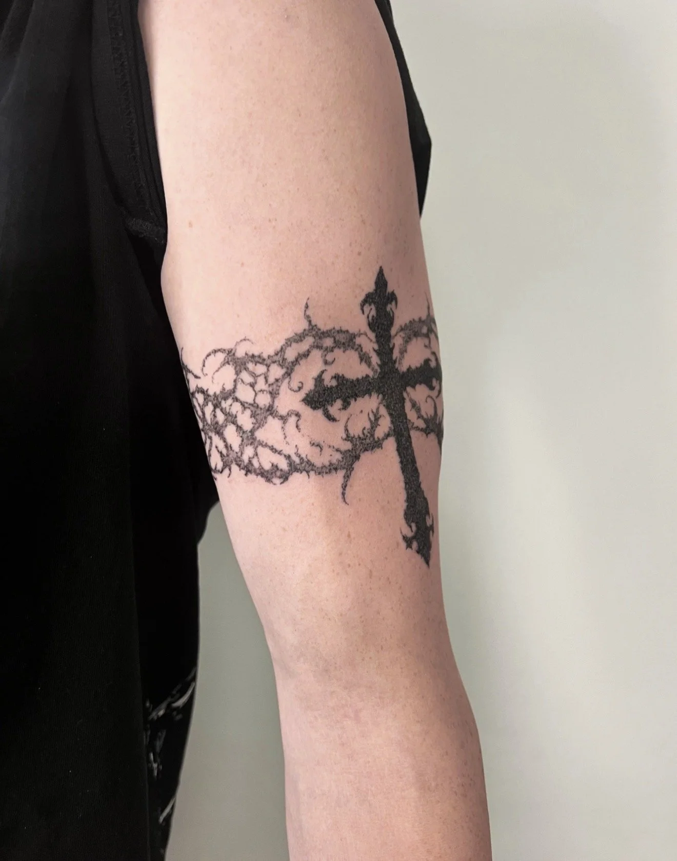 Tattoo of a large black cross with intricate, thorny wings on a person's upper arm.