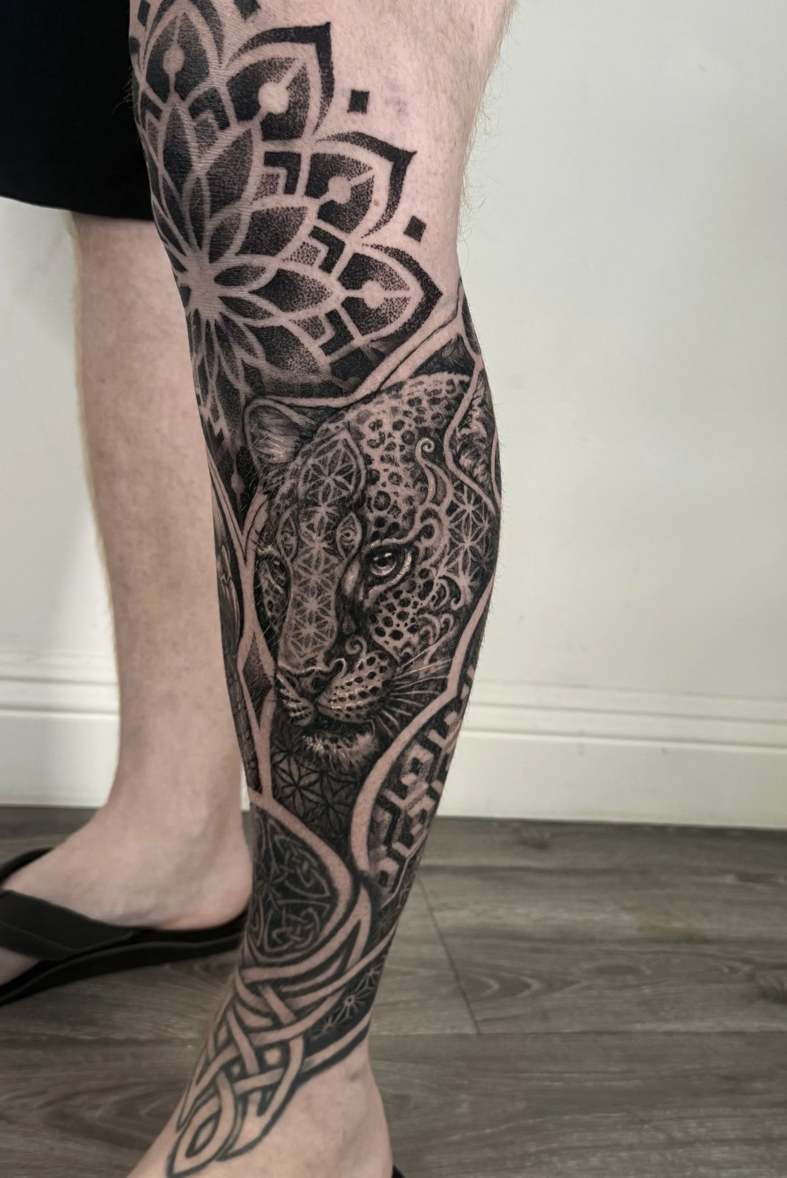 Tattoo of a leopard's face surrounded by geometric patterns on a person's thigh and calf.
