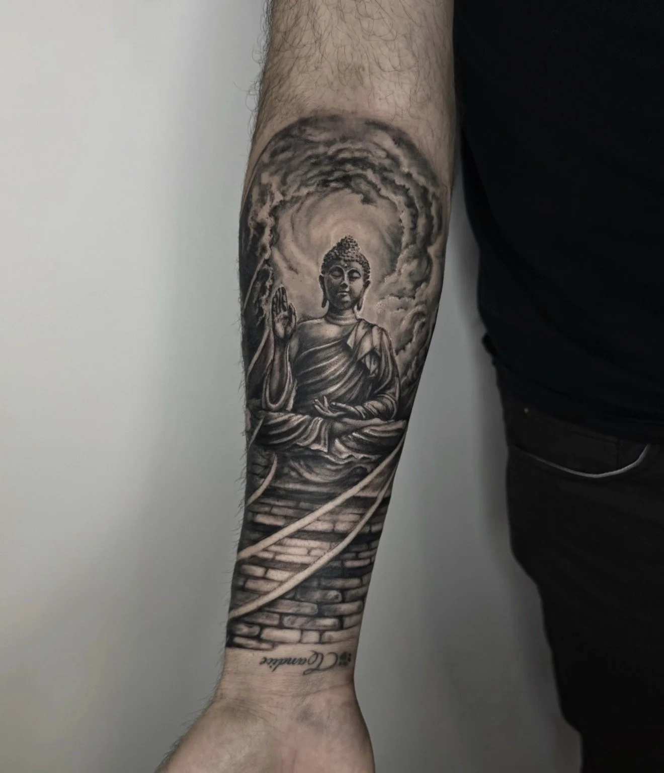 Black and gray tattoo of Buddha sitting cross-legged with hand raised in blessing, surrounded by clouds and a staircase leading up to him.