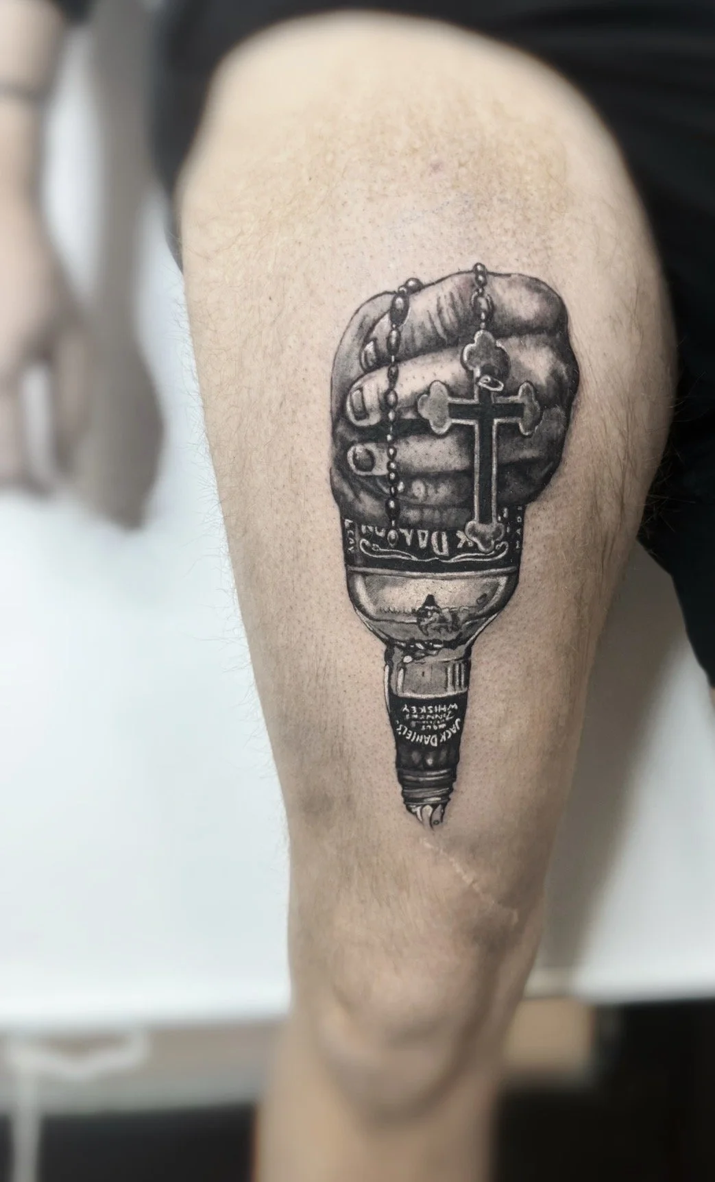 Black and gray tattoo on person's thigh depicting a hand holding a rosary and a beer bottle, with a cross and text on the bottle.