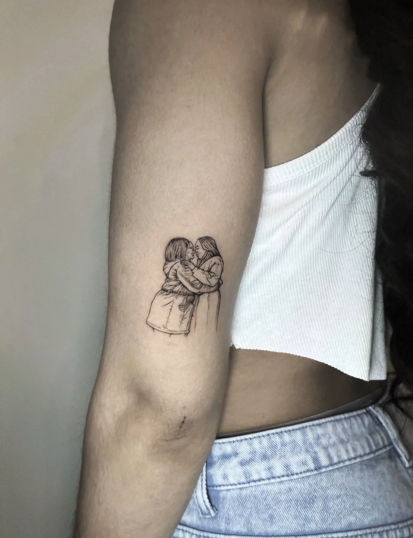 A person with a tattoo of two women embracing, one with short hair and the other with long hair, on their upper arm. The person is wearing a white sleeveless top and blue jeans.