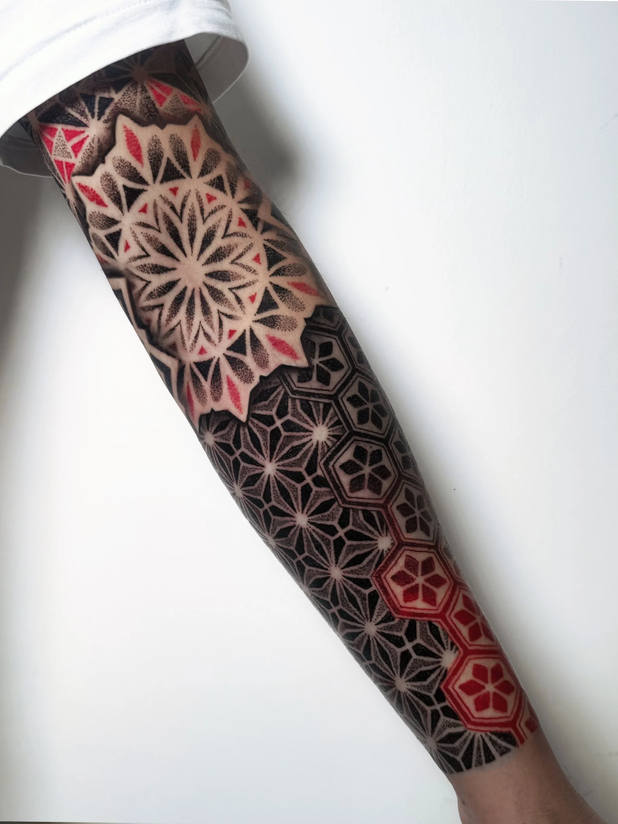 Forearm tattoo with geometric patterns, featuring large starburst design in cream, black, red, and gray, with some black hexagons and red lines.