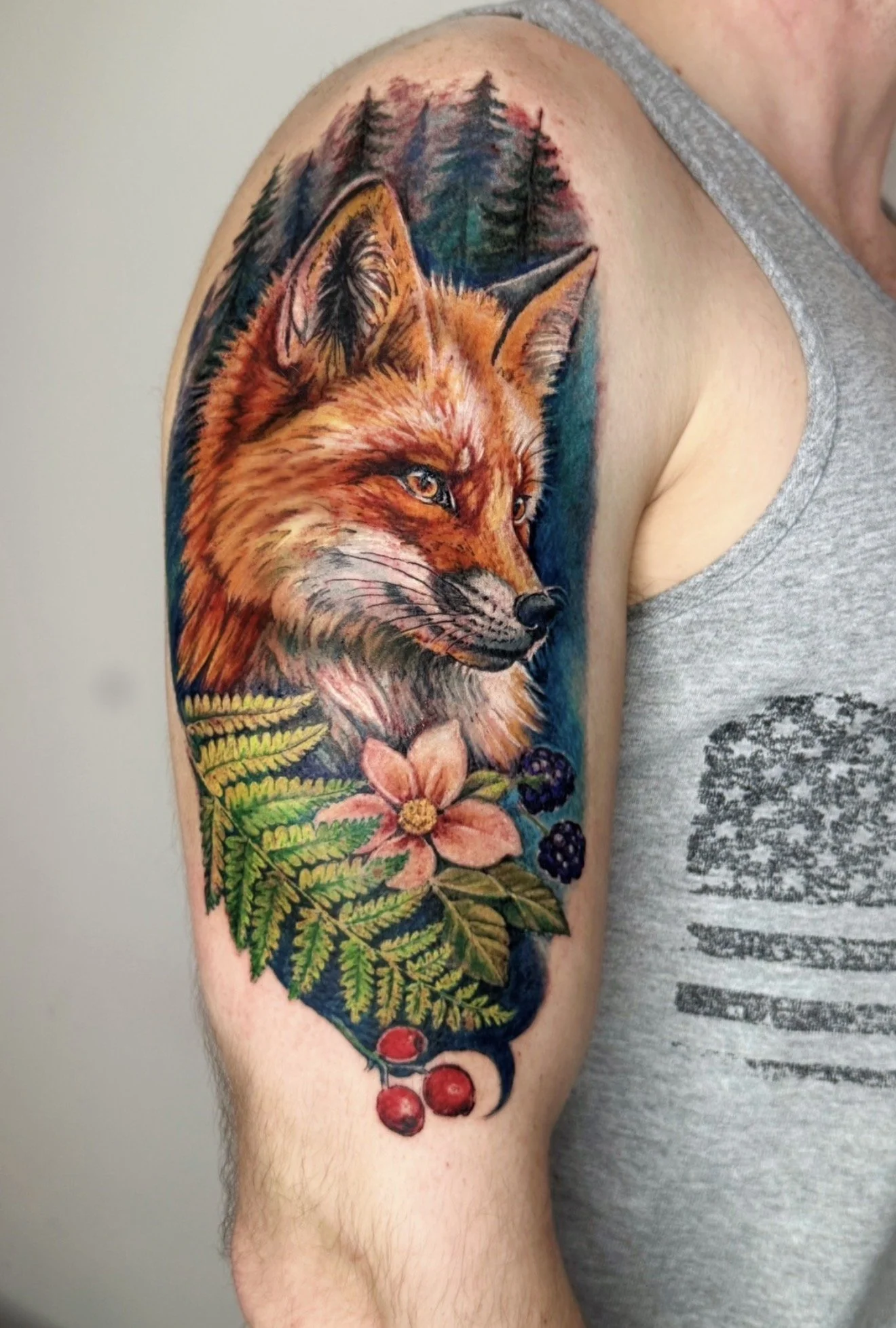 Colorful tattoo of a fox surrounded by pine trees, flowers, and berries on a person's upper arm.