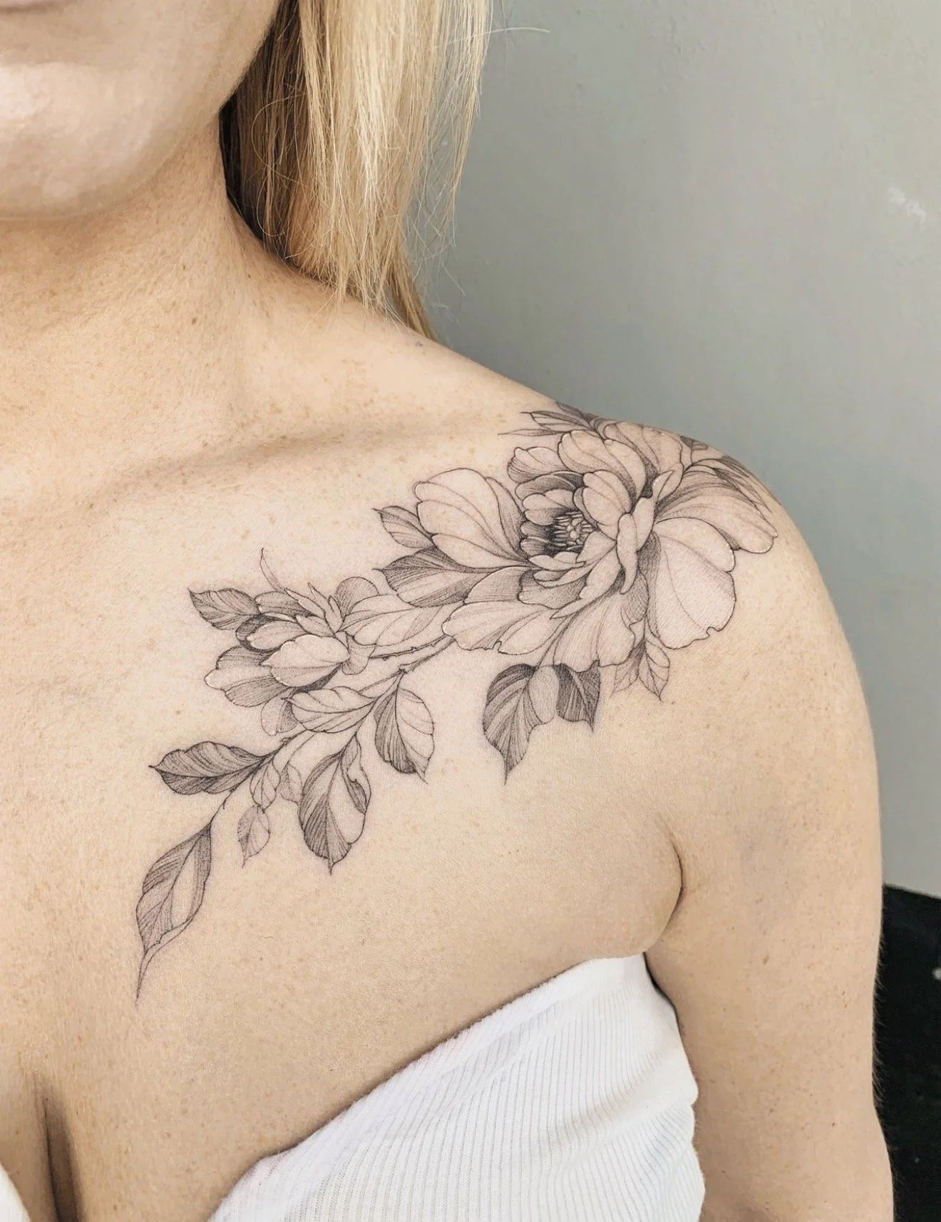Close-up of a person's shoulder and chest with a floral tattoo that features a large flower and leaves. The person is wearing a white strapless garment.