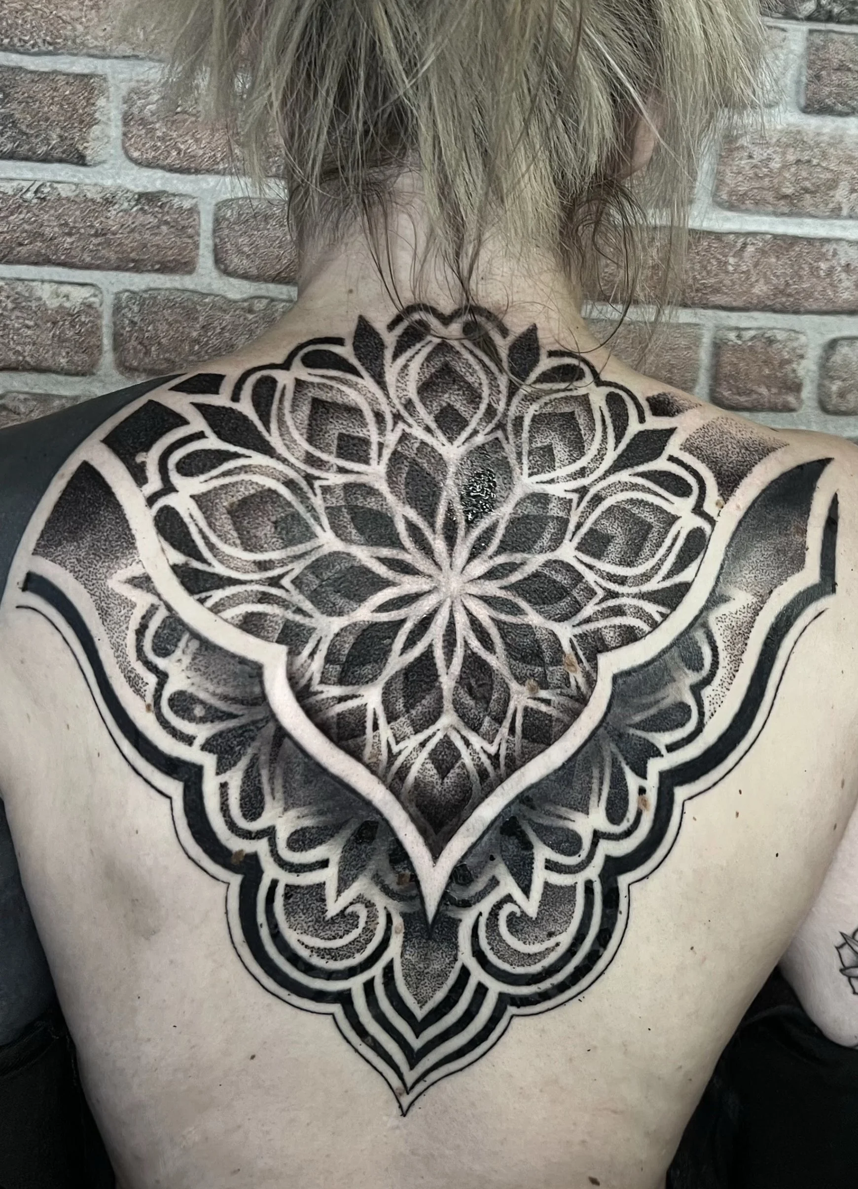 A person's upper back with an intricate black and white geometric tattoo covering the shoulder and upper chest area. The tattoo features symmetrical patterns and floral elements, with the person's hair hanging down over the neck.