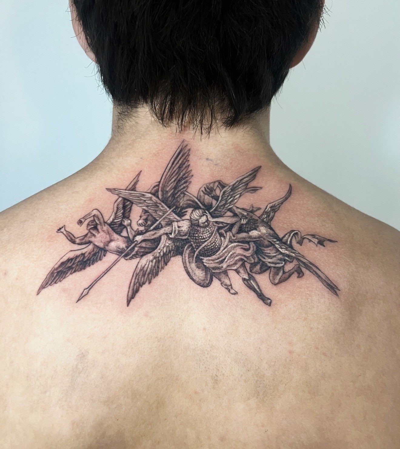 A person with short dark hair shows the back of their neck and upper shoulders, featuring a detailed black and gray tattoo of a large insect with multiple wings and a human figure riding it.