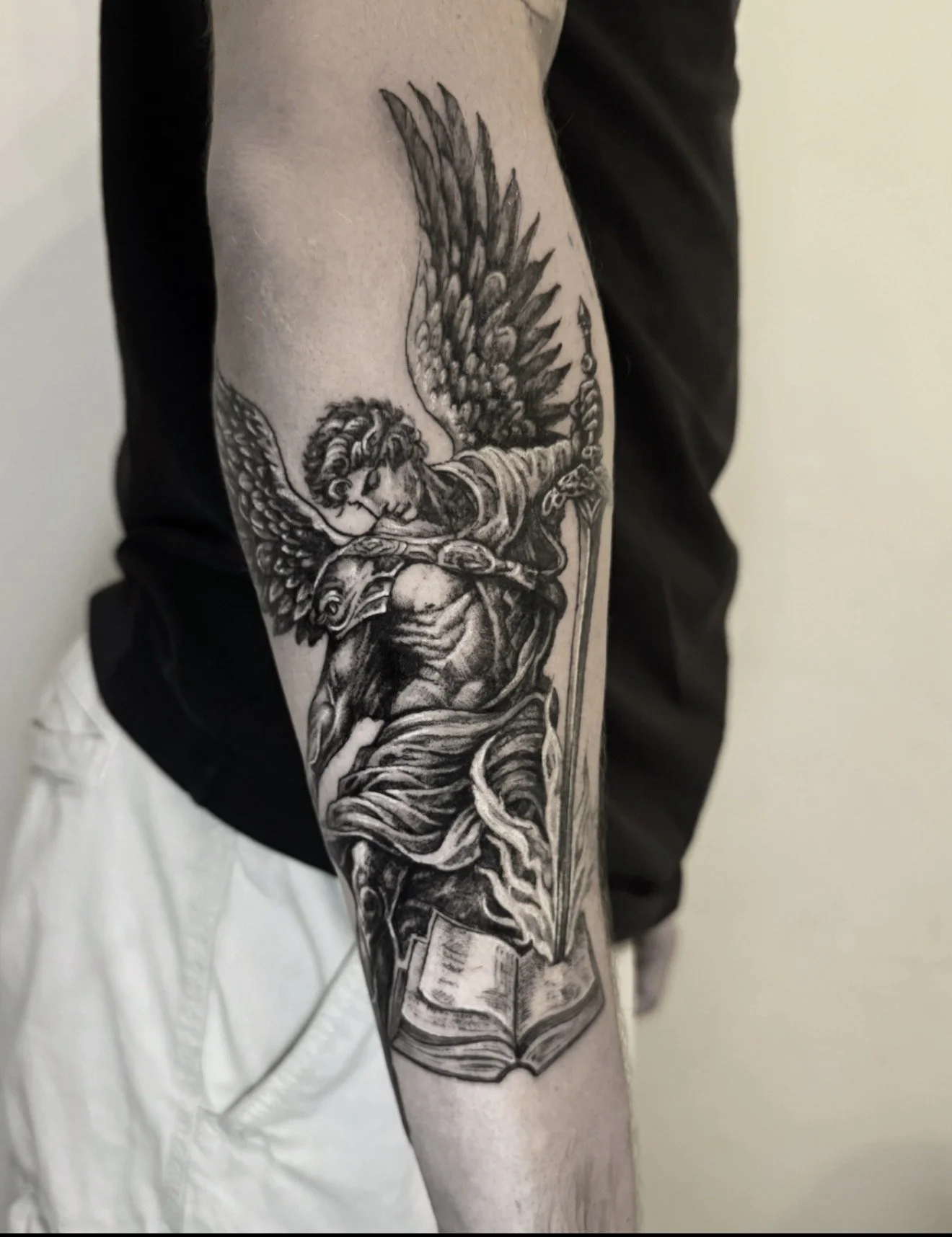 Black and gray tattoo on person's arm of a winged angel holding a sword, with an open book at the bottom.
