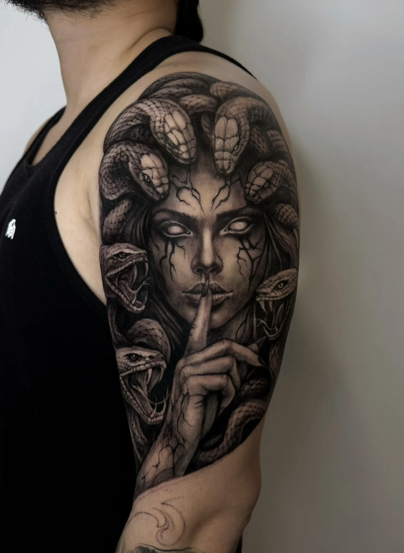 Black and gray tattoo of a woman's face with snakes around her head and hand, and a finger held to her lips, on a person's upper arm and shoulder.