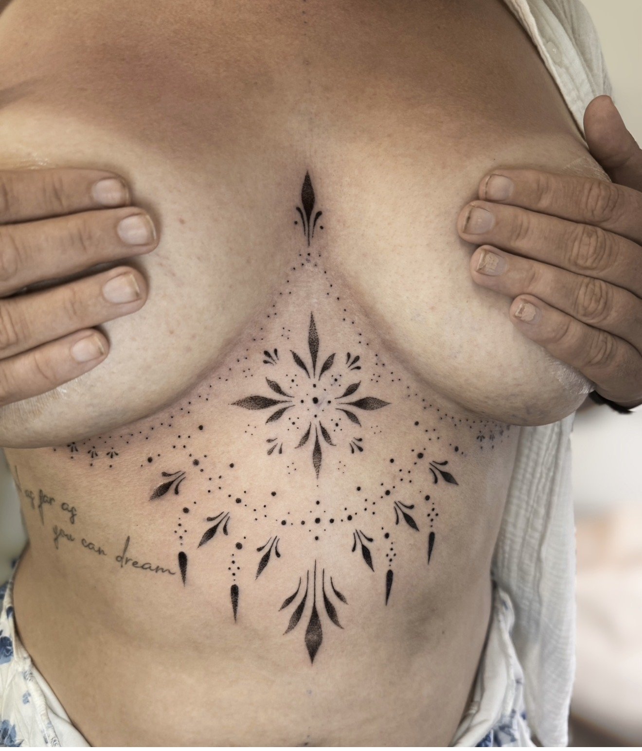 A person with intricate black tattoos on their chest and abdomen, partially covering their breasts with hands.
