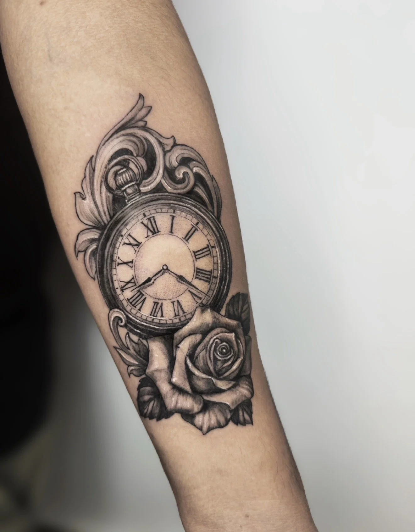 Black and gray tattoo of a pocket watch with Roman numerals, surrounded by detailed roses and decorative scrollwork, on a person's forearm.