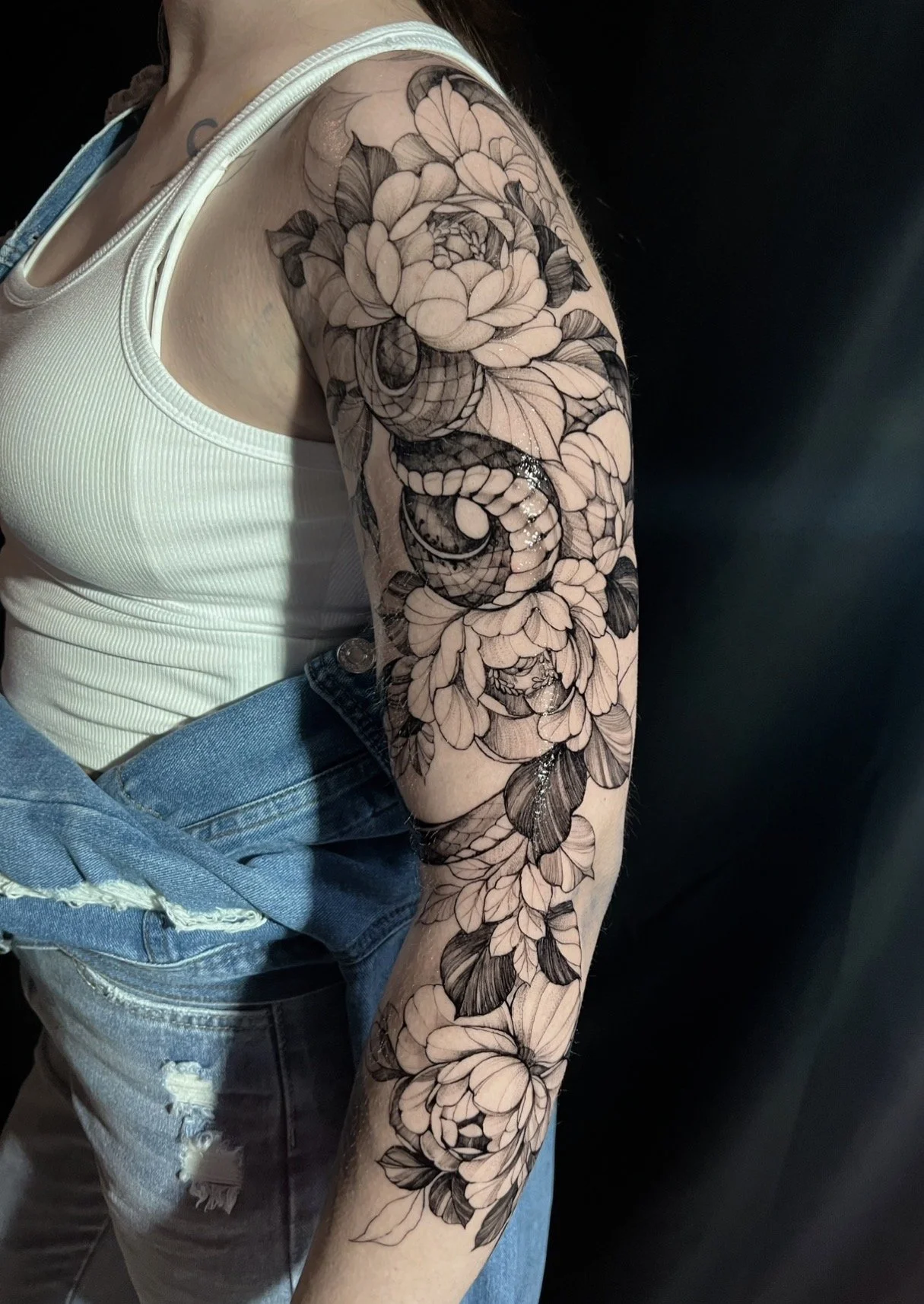 A black and gray floral tattoo sleeve with peonies, leaves, and a snake, worn on a woman's arm.