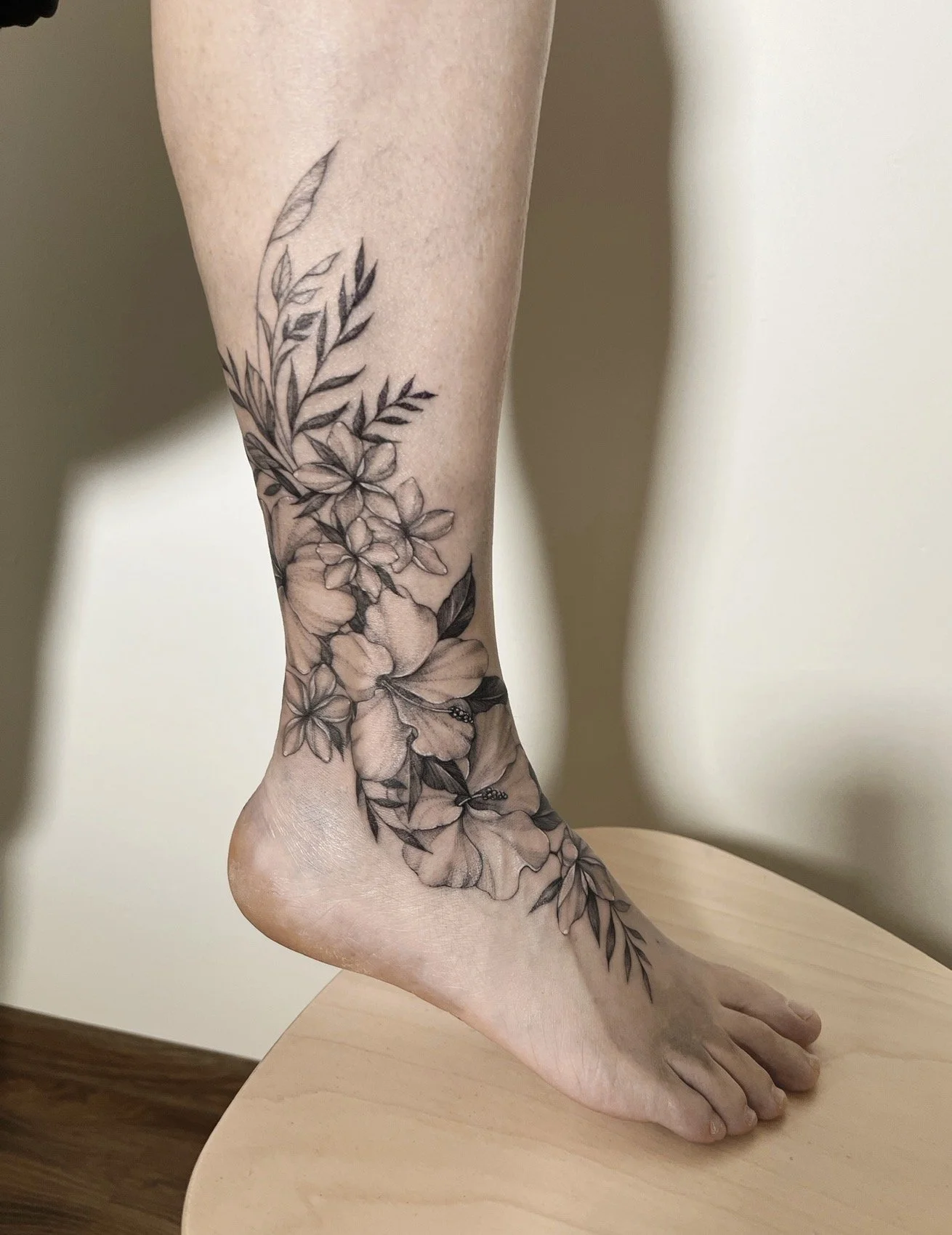 Black and gray floral tattoo on person's foot and ankle, featuring various flowers and leaves.