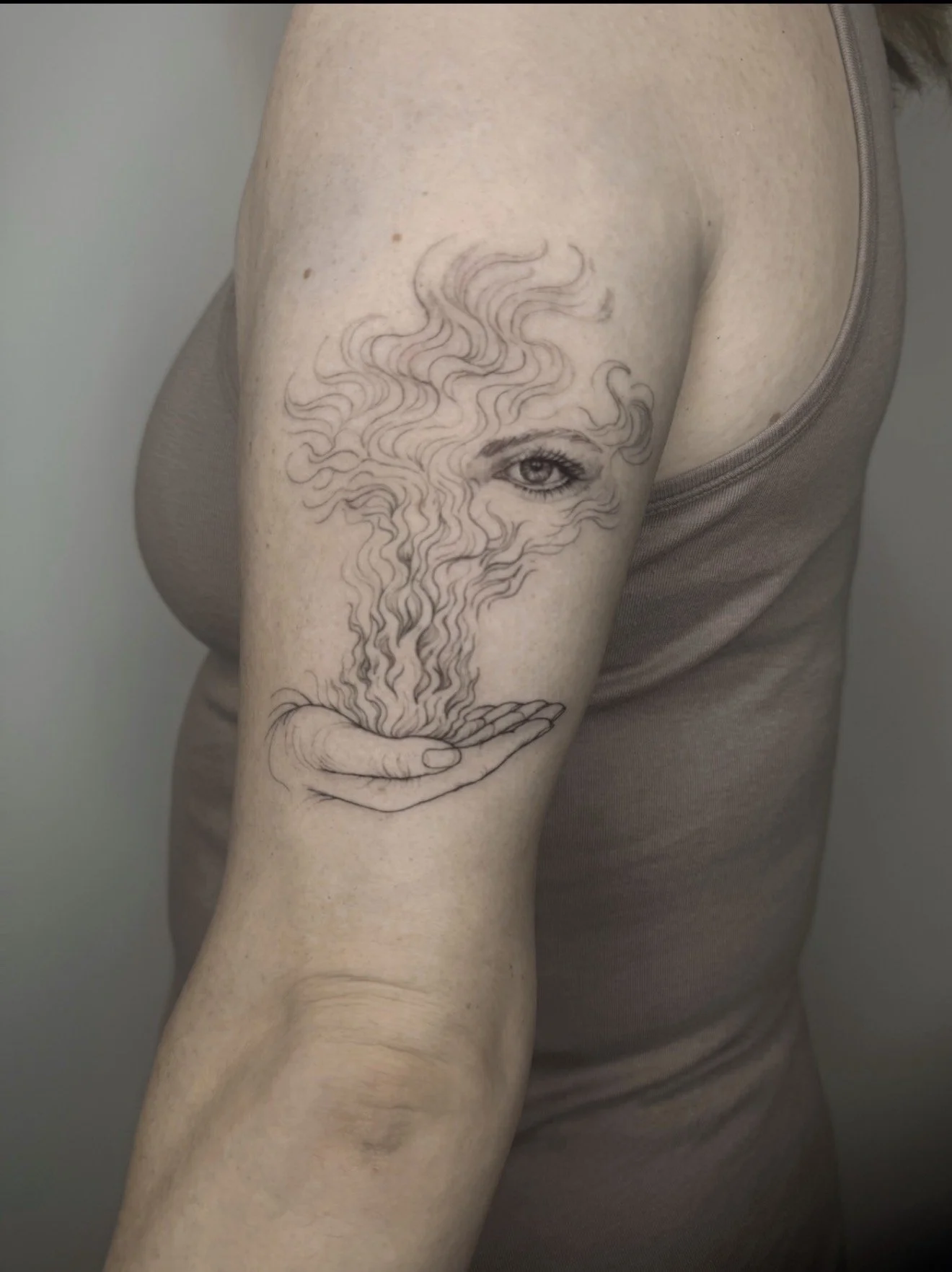 Tattoo of a woman's face with flowing hair, held in a hand, on the upper arm of a person wearing a tank top.
