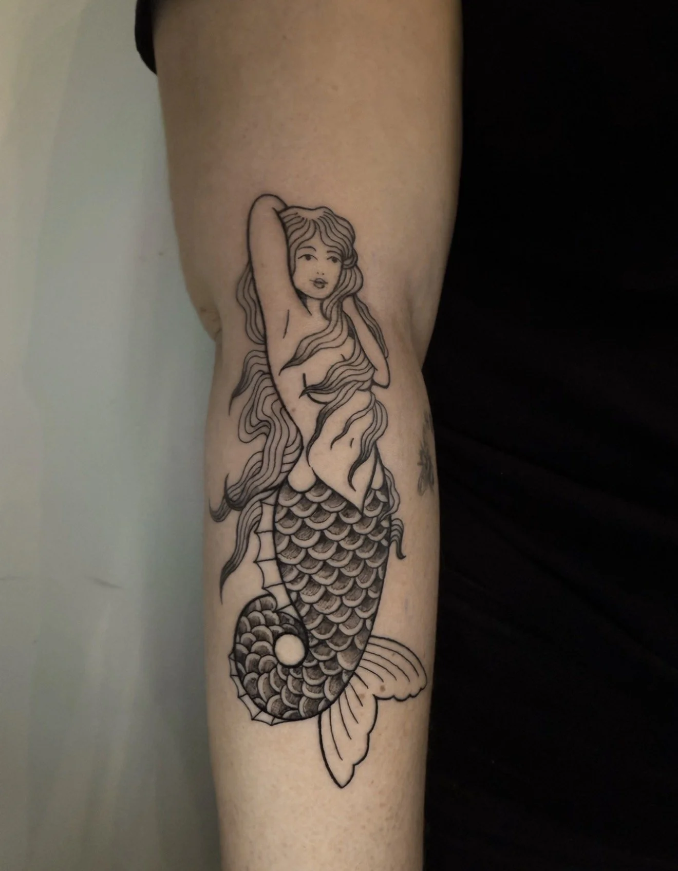 Tattoo of a mermaid with long hair and a fish tail, depicted with a cartoon style, on a person's arm.