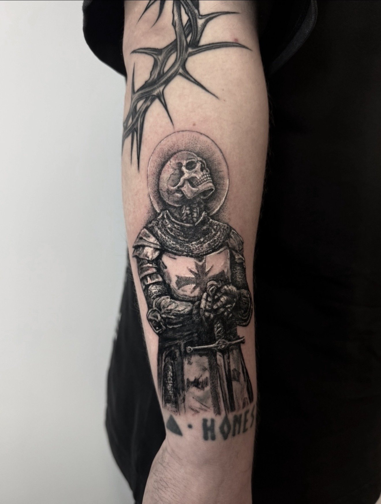 Black and gray tattoo of a skeleton in armor with a halo behind its head, holding a sword, on a person's arm.