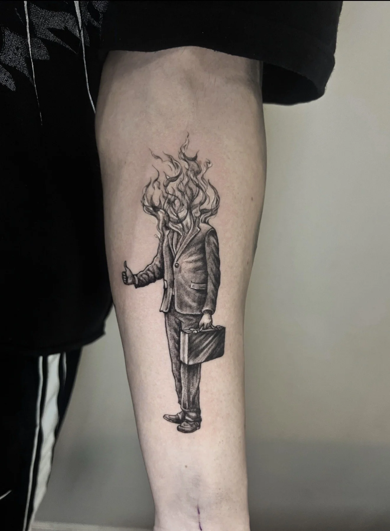 Tattoo of a man in a suit with the head transformed into flames, holding a briefcase in one hand and giving a thumbs-up with the other, on a person's leg.