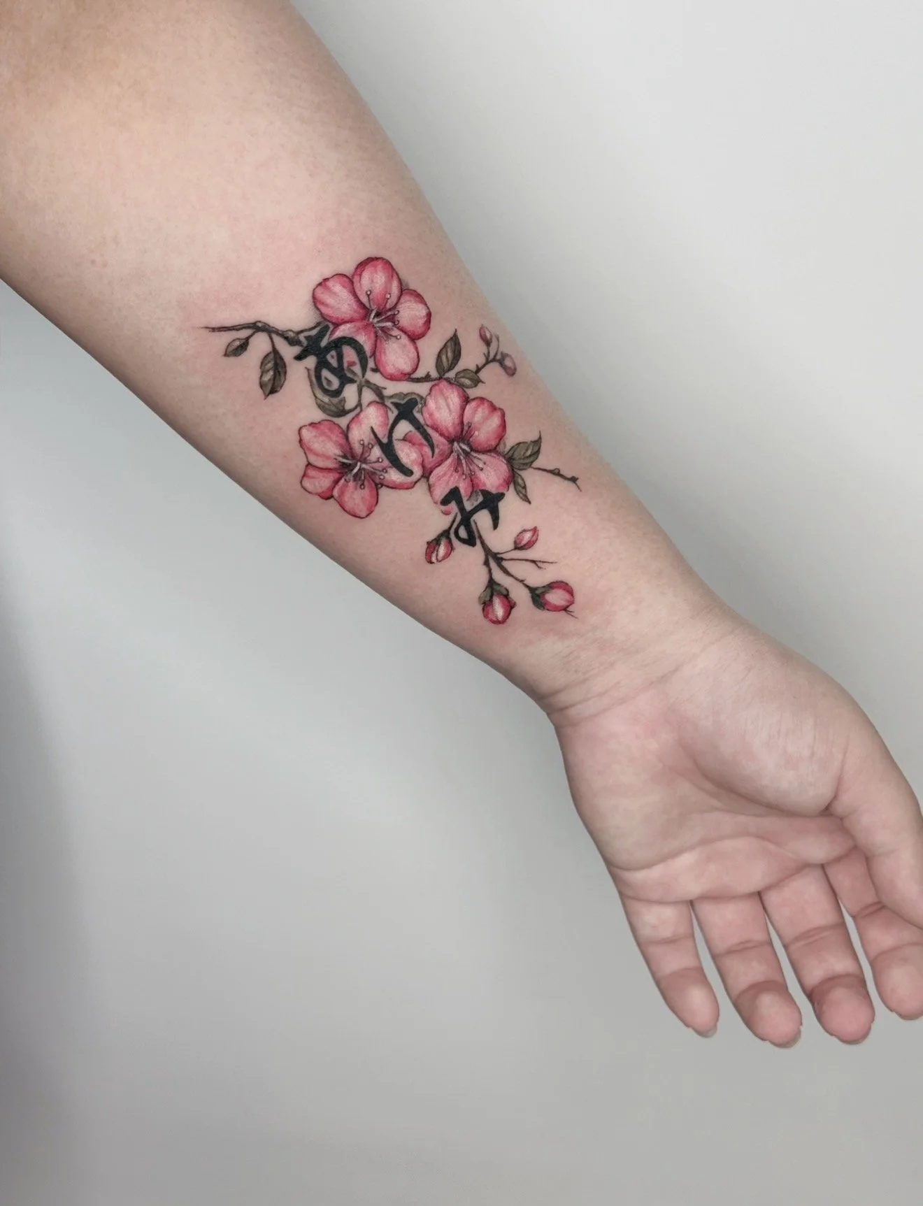Colorful tattoo of pink cherry blossoms on a person's forearm with Japanese characters integrated within the floral design.