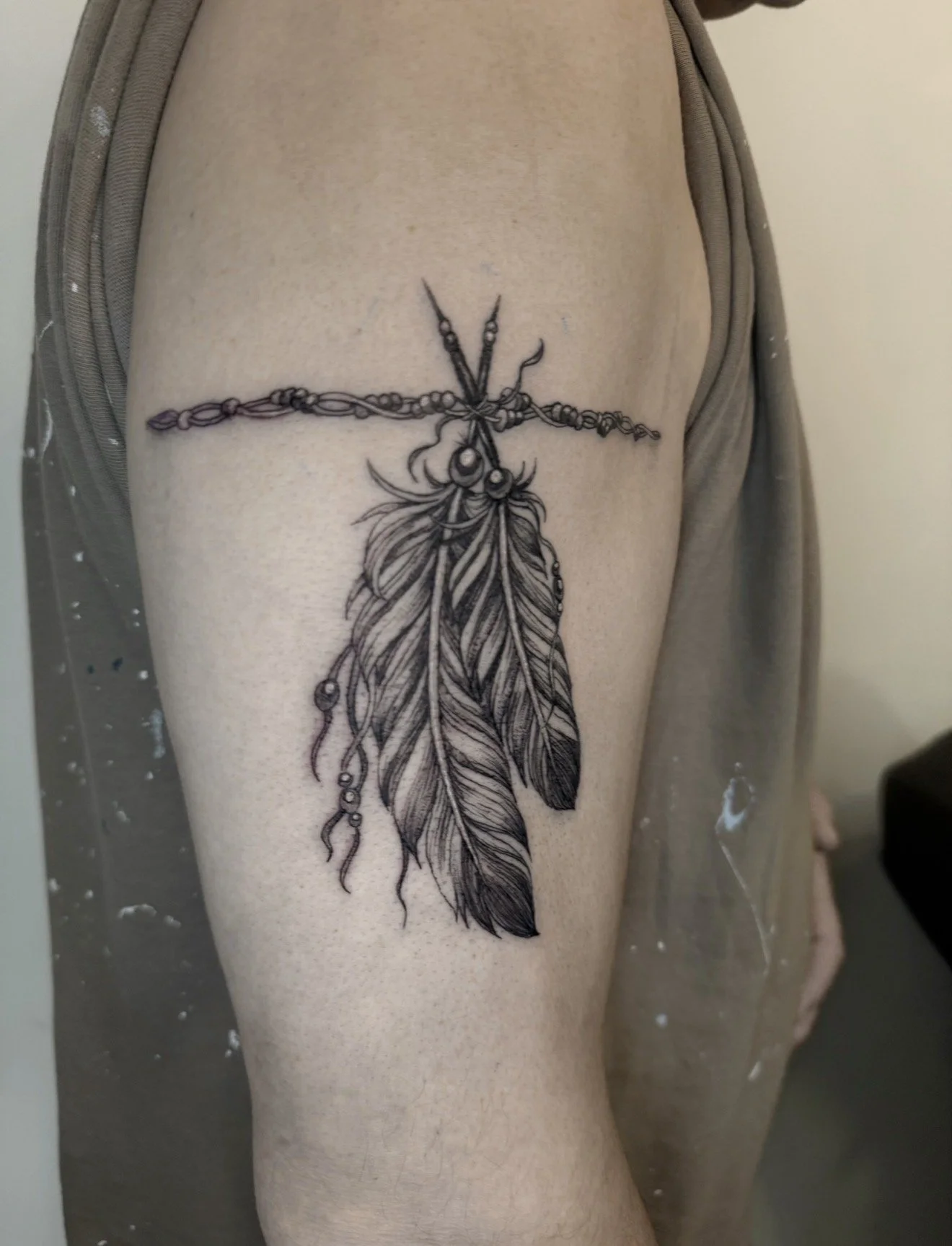 A tattoo on an upper arm of a cross made of feathers and beads, with a large feather hanging below.