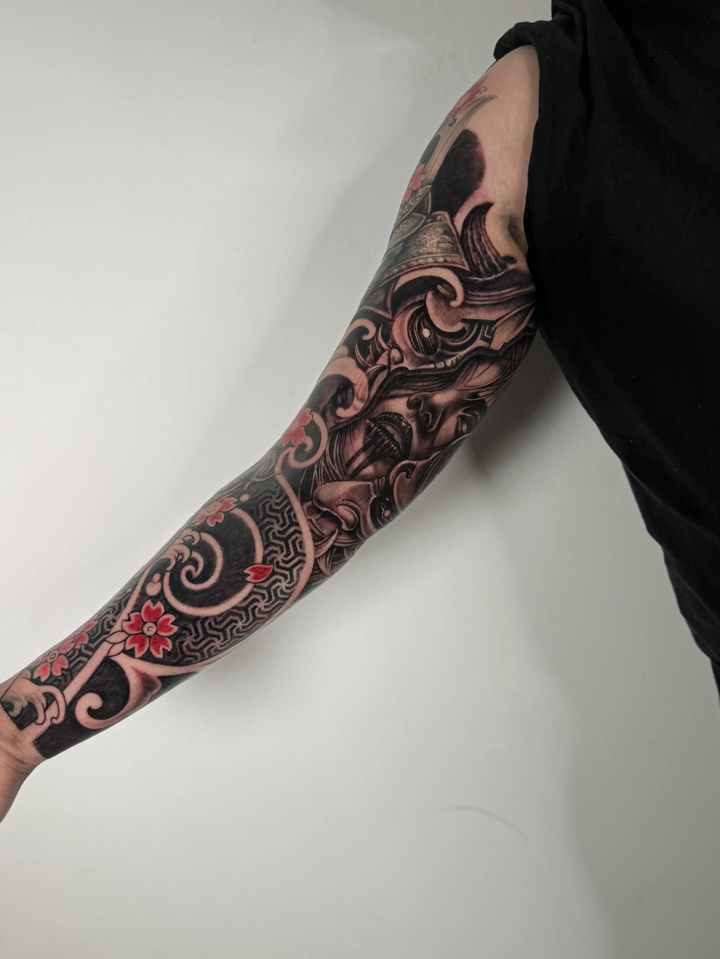 Close-up of a person's arm with intricate Japanese-style tattoos, including waves, cherry blossoms, and traditional motifs, against a plain white background. The person is wearing a black short-sleeved shirt.