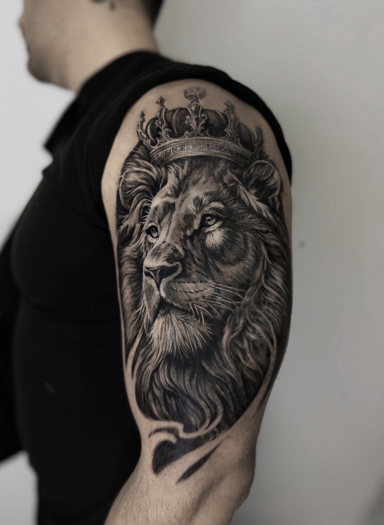 Black and gray tattoo of a lion's head with a crown on its head, on a person's upper arm.