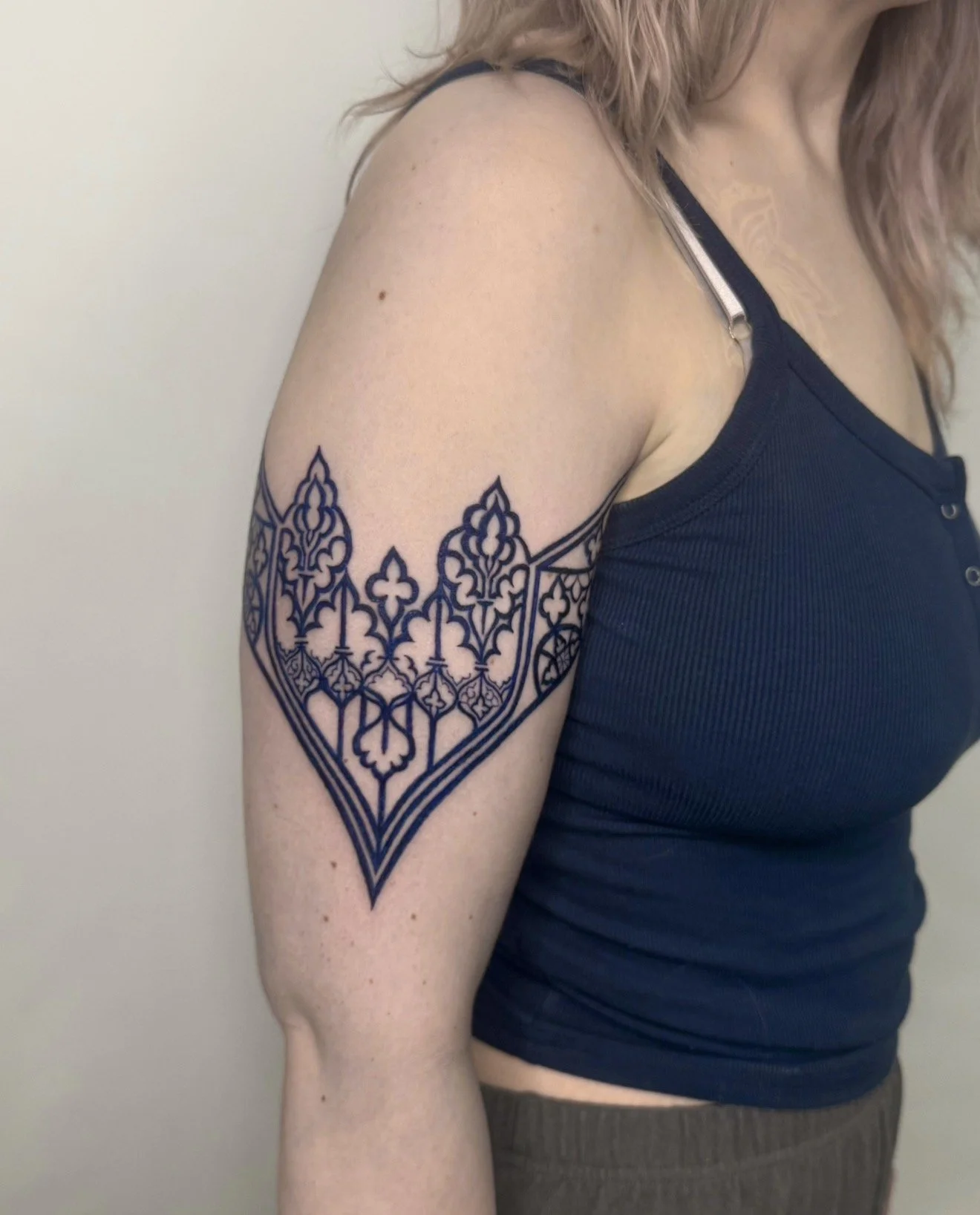 Close-up of a woman's upper arm showing a large, intricate, dark blue tattoo with geometric and floral patterns. She is wearing a dark tank top with a metal strap and stands against a plain, off-white background.