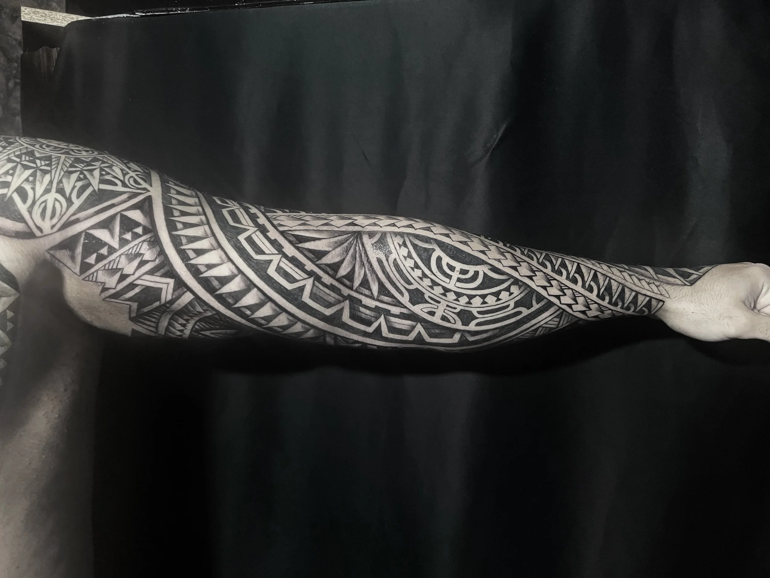 A person with black and white patterned tattooed arm and leg sitting on a dark surface, possibly a couch or bed.