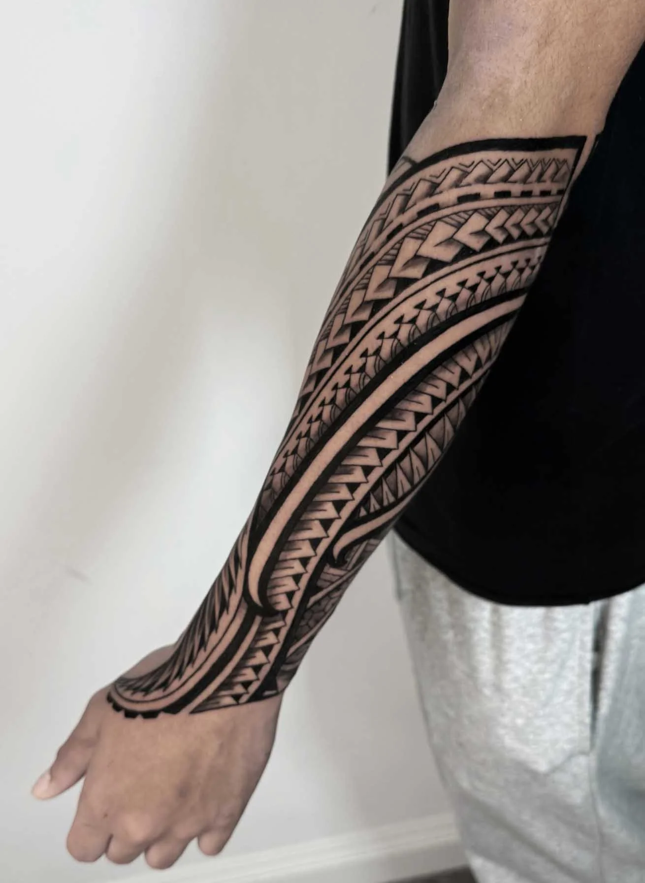 A person's arm with an intricate black Polynesian-style tattoo covering from the shoulder to the wrist, featuring geometric patterns and lines.