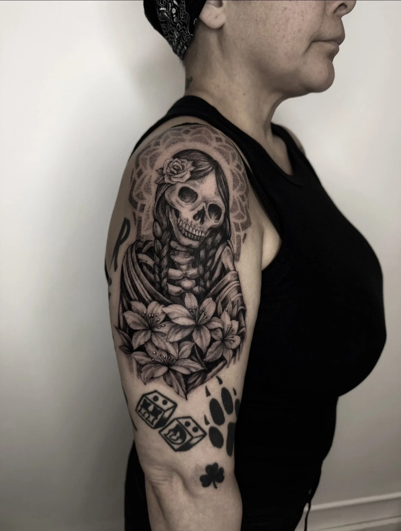 A woman with a black bandana and black top showing her left arm and shoulder tattoo of a skull with a woman's face, flowers, and geometric patterns.