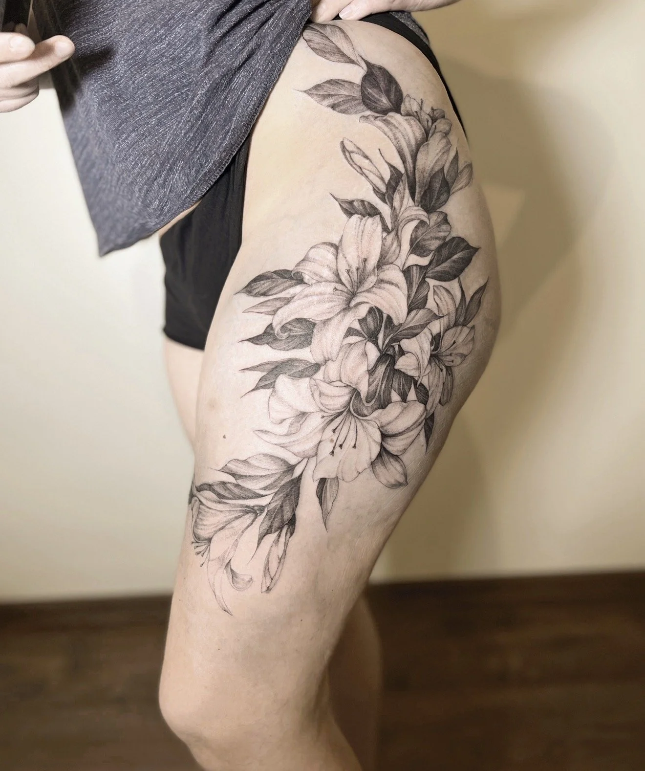 A person's upper thigh and hip featuring a detailed black and gray floral tattoo with large lilies and leaves.