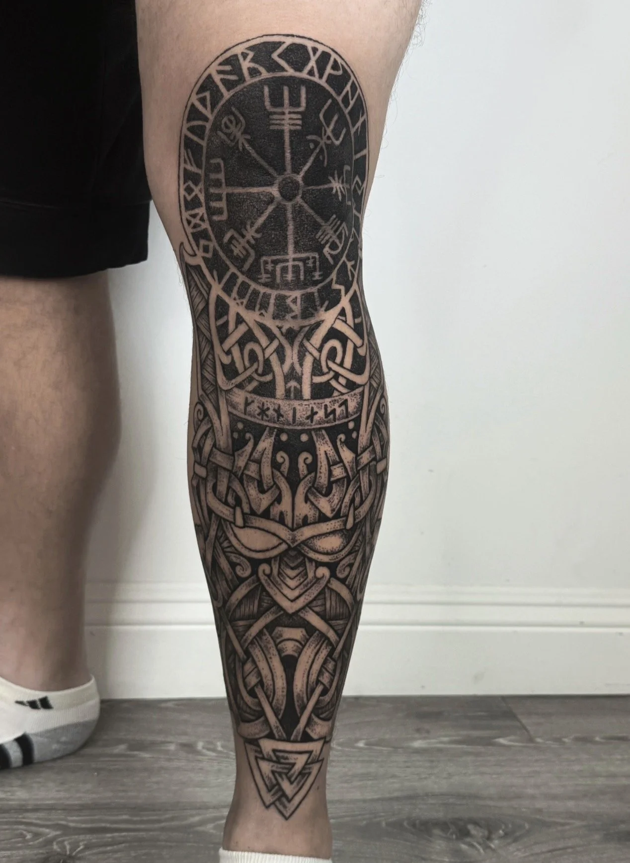 A person's leg with a detailed black and gray tattoo sleeve featuring patterns inspired by Norse and Celtic designs, including runes, knot work, and a central mystical symbol.