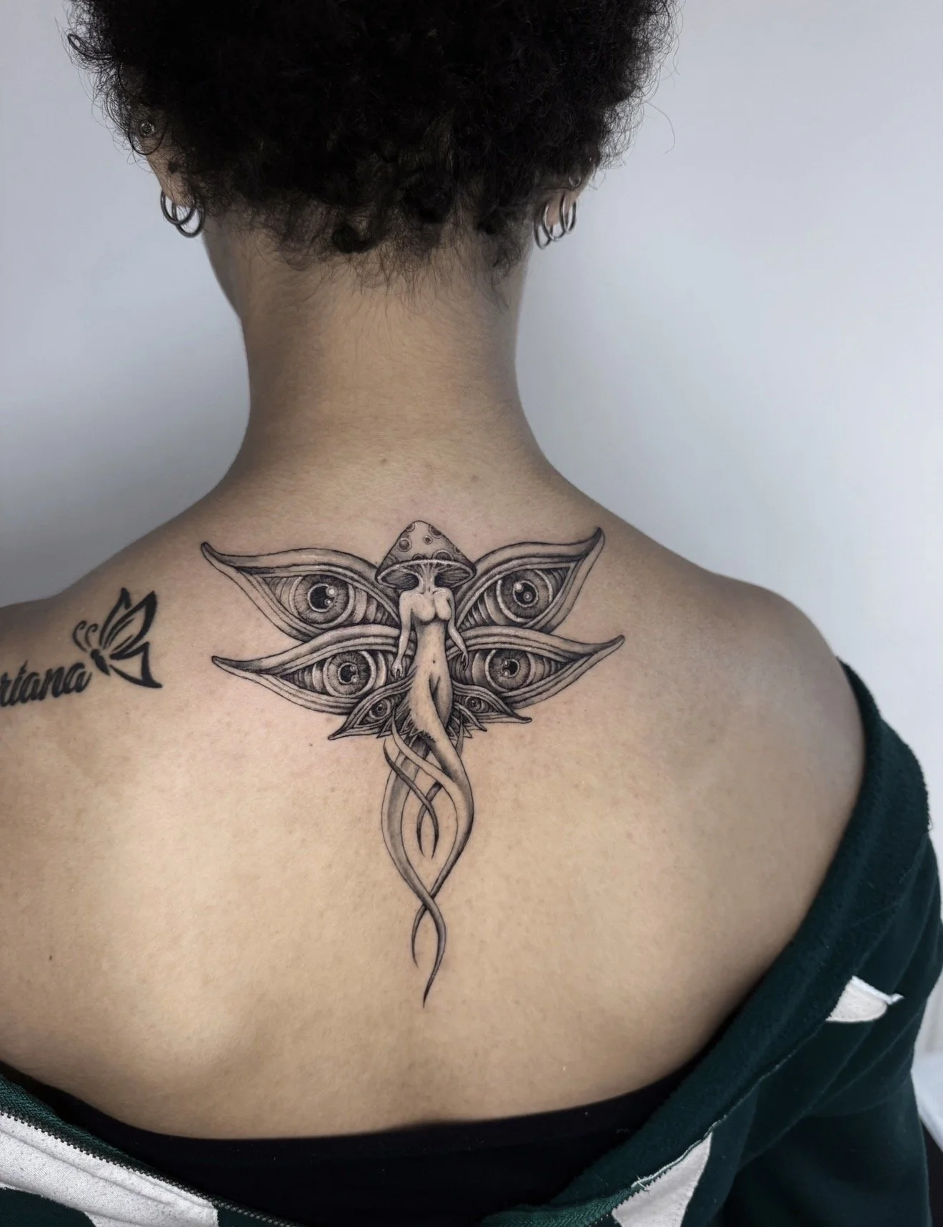 Back view of a person with short curly hair, wearing earrings, showing a detailed black and gray tattoo on the upper back and shoulder. The tattoo features a humanoid figure with butterfly wings that have numerous eyes, and a snake intertwined below.