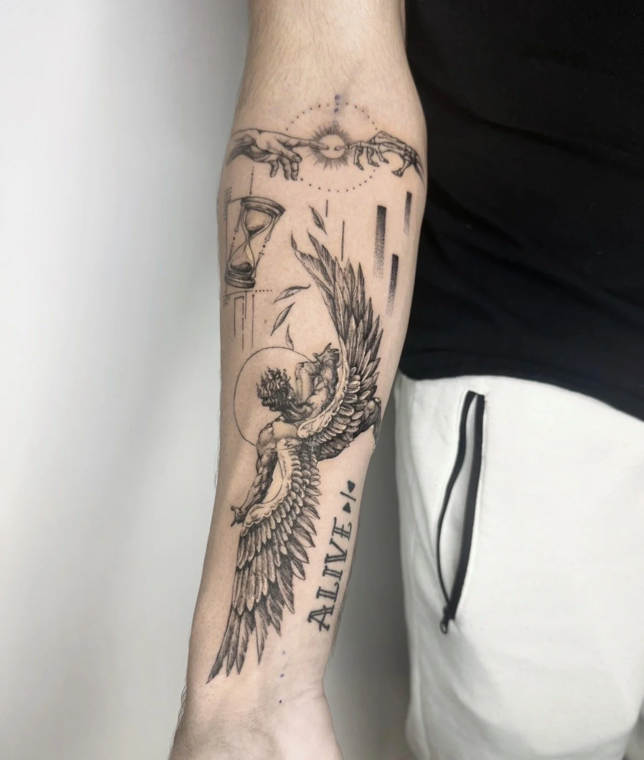 A tattoo on a person's forearm featuring a detailed angel with wings, a muscular figure, and the word 'ALIVE' written vertically. Above, there are geometric and abstract elements, including a sun, and hands reaching toward each other.