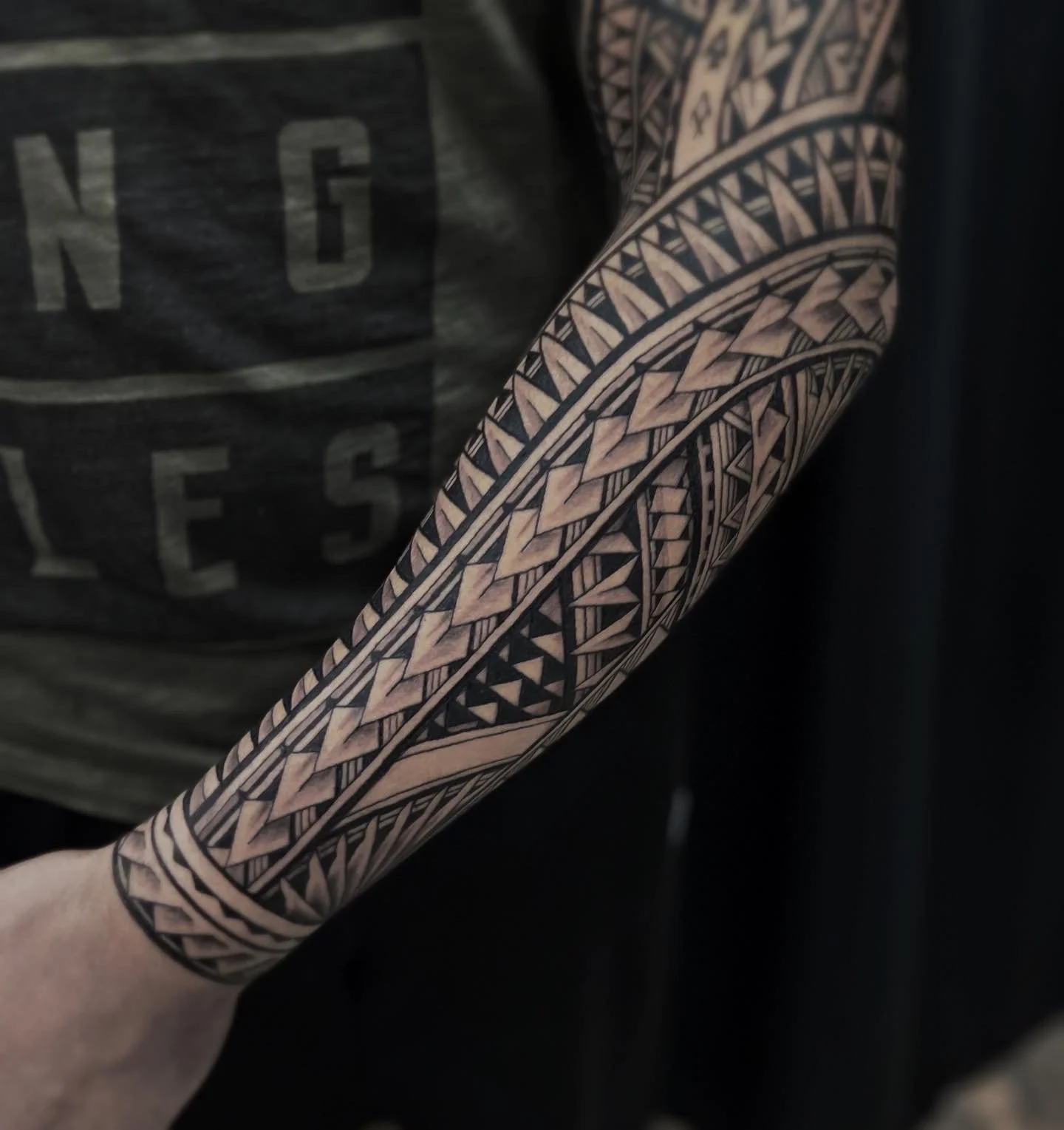 Close-up of a person's arm with an intricate black and beige geometric tattoo sleeve, adjacent to a black shirt with faded text.