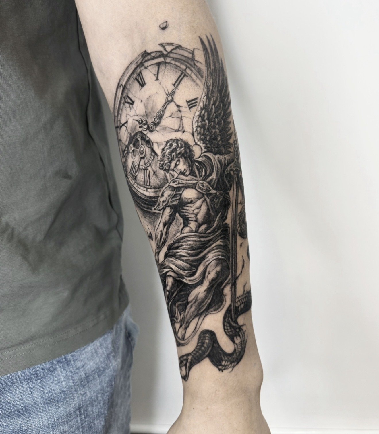 Black and gray tattoo of a classical angel with wings, holding a sword, with a broken clock in the background on a person's forearm.