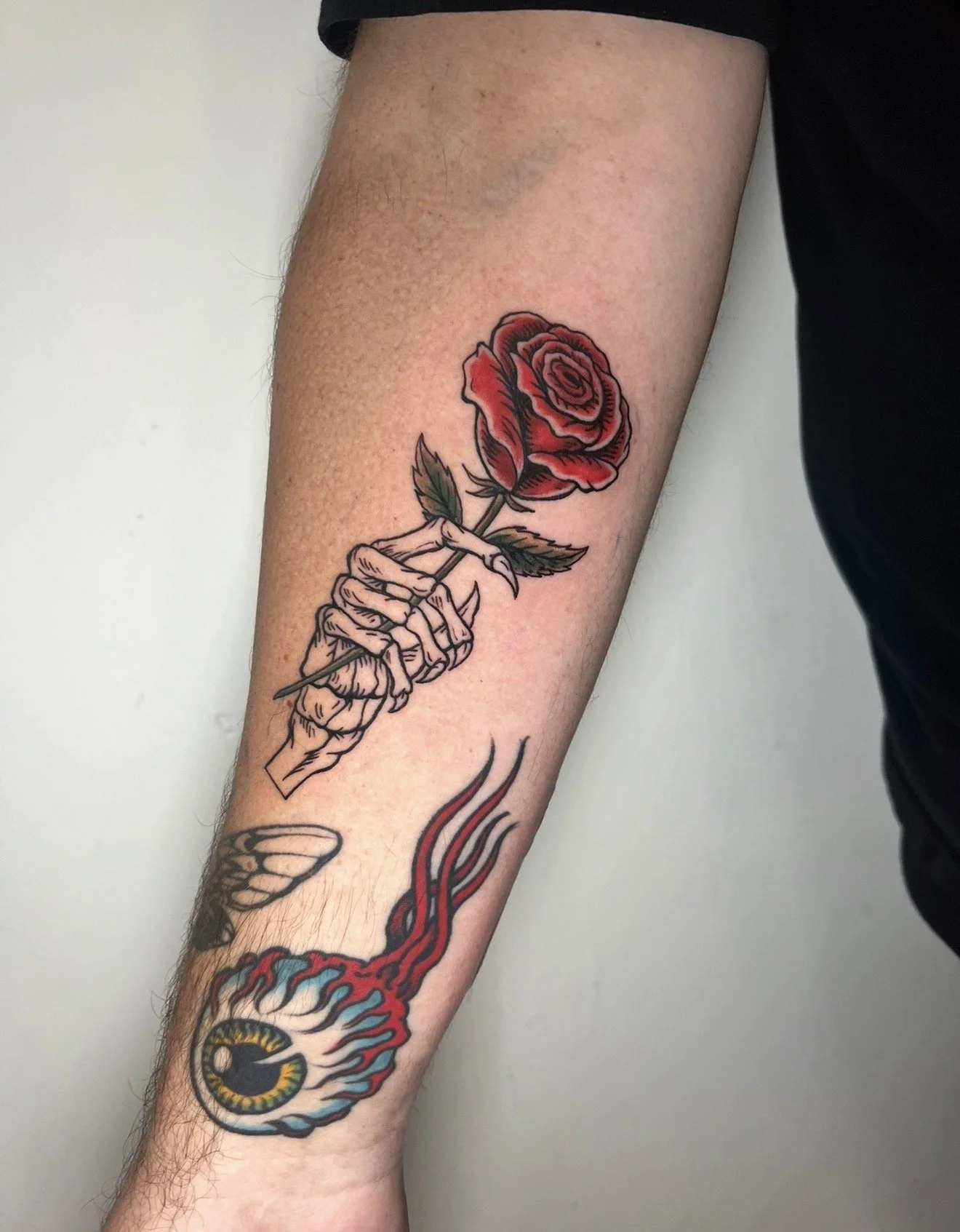 Tattoo of a skeletal hand holding a red rose with green leaves. The tattoo is on a person's forearm.