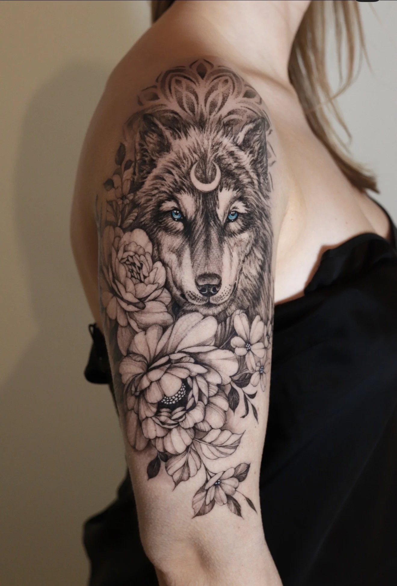 A detailed black and gray tattoo of a wolf with bright blue eyes, surrounded by flowers, on a person's upper arm and shoulder.