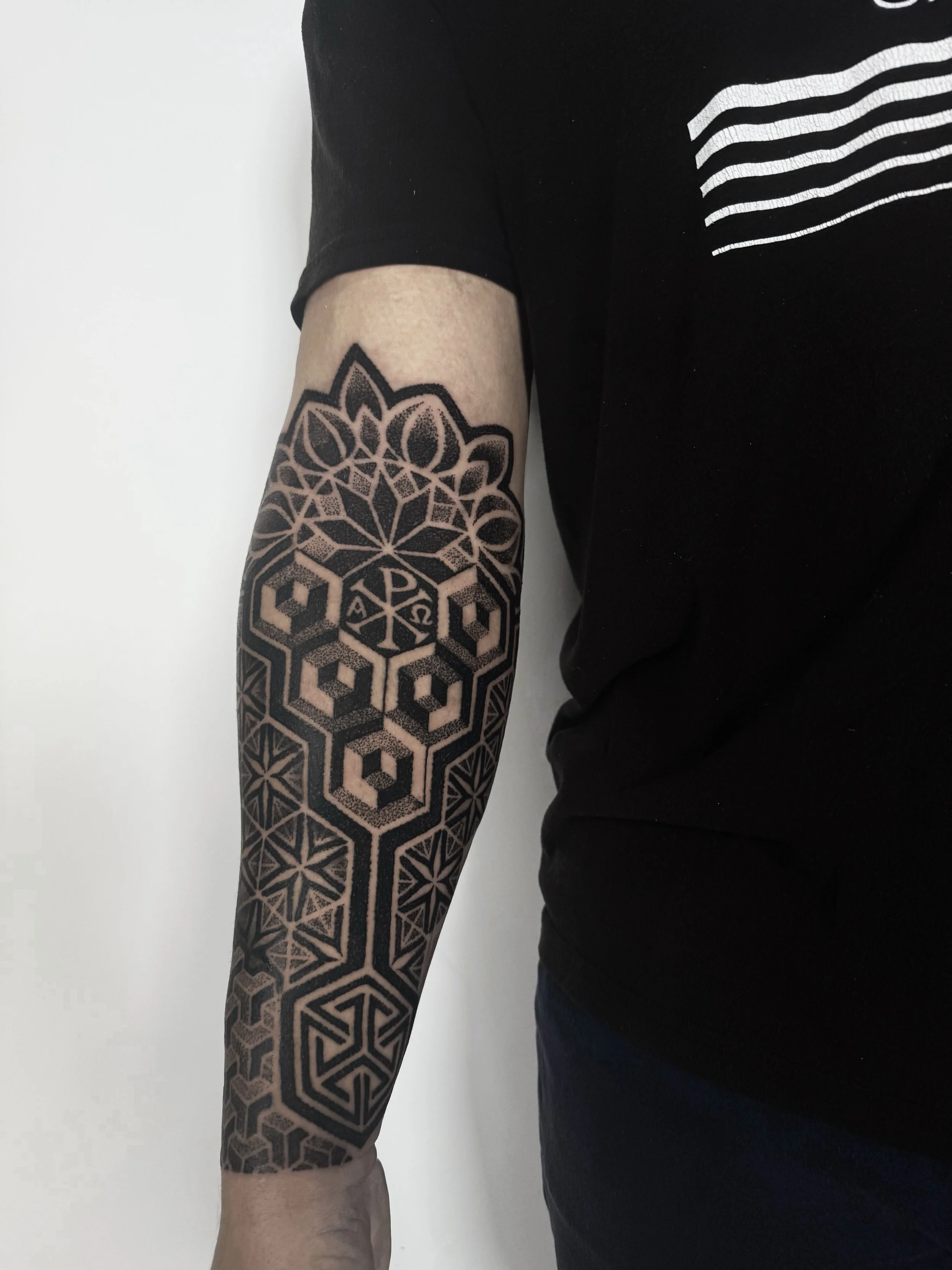 A person's arm with an intricate black tattoo sleeve featuring geometric patterns and Christian symbols, wearing a black T-shirt with white stripes.