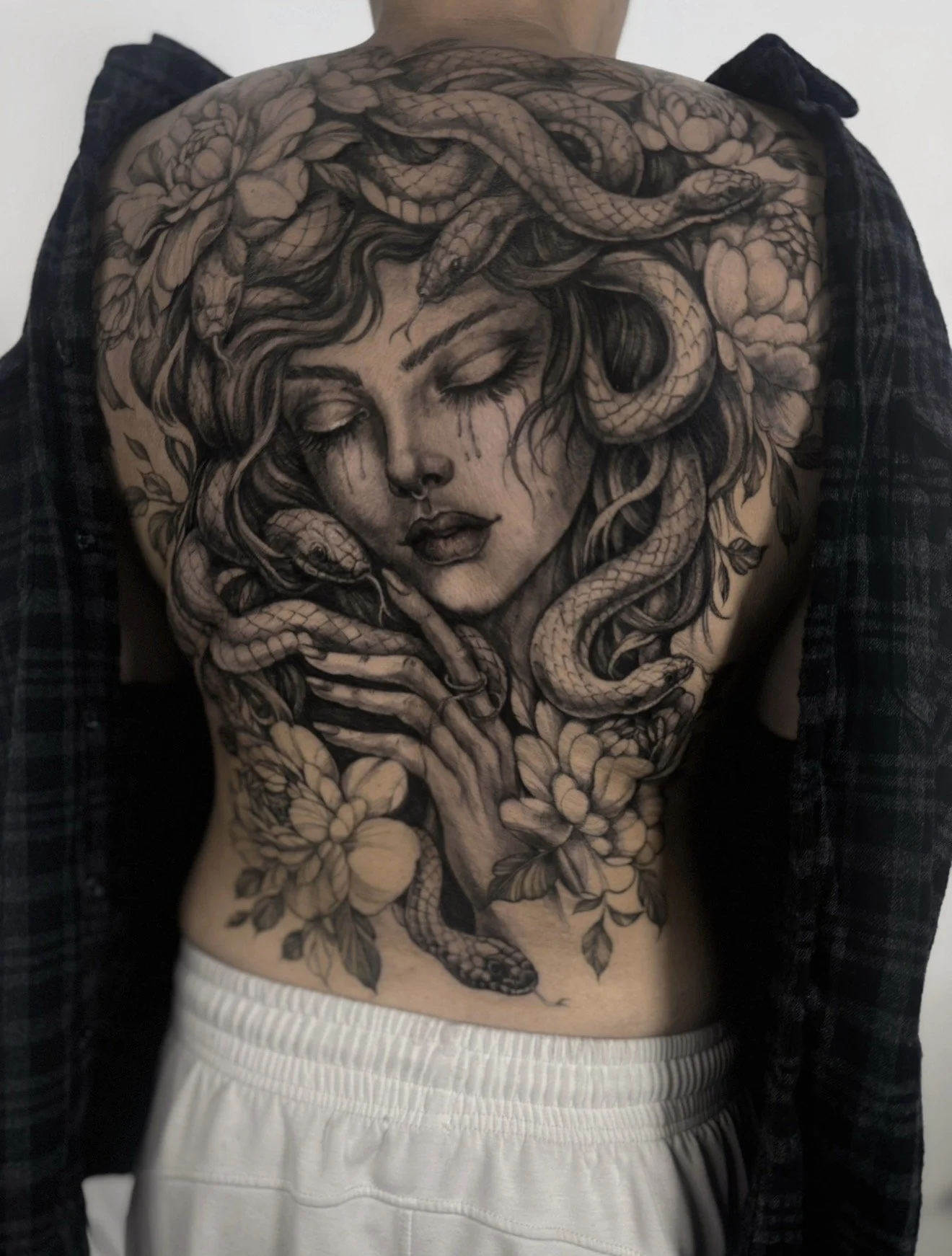 Black and gray tattoo of a woman's face with closed eyes, surrounded by snakes and florals on a person's back.