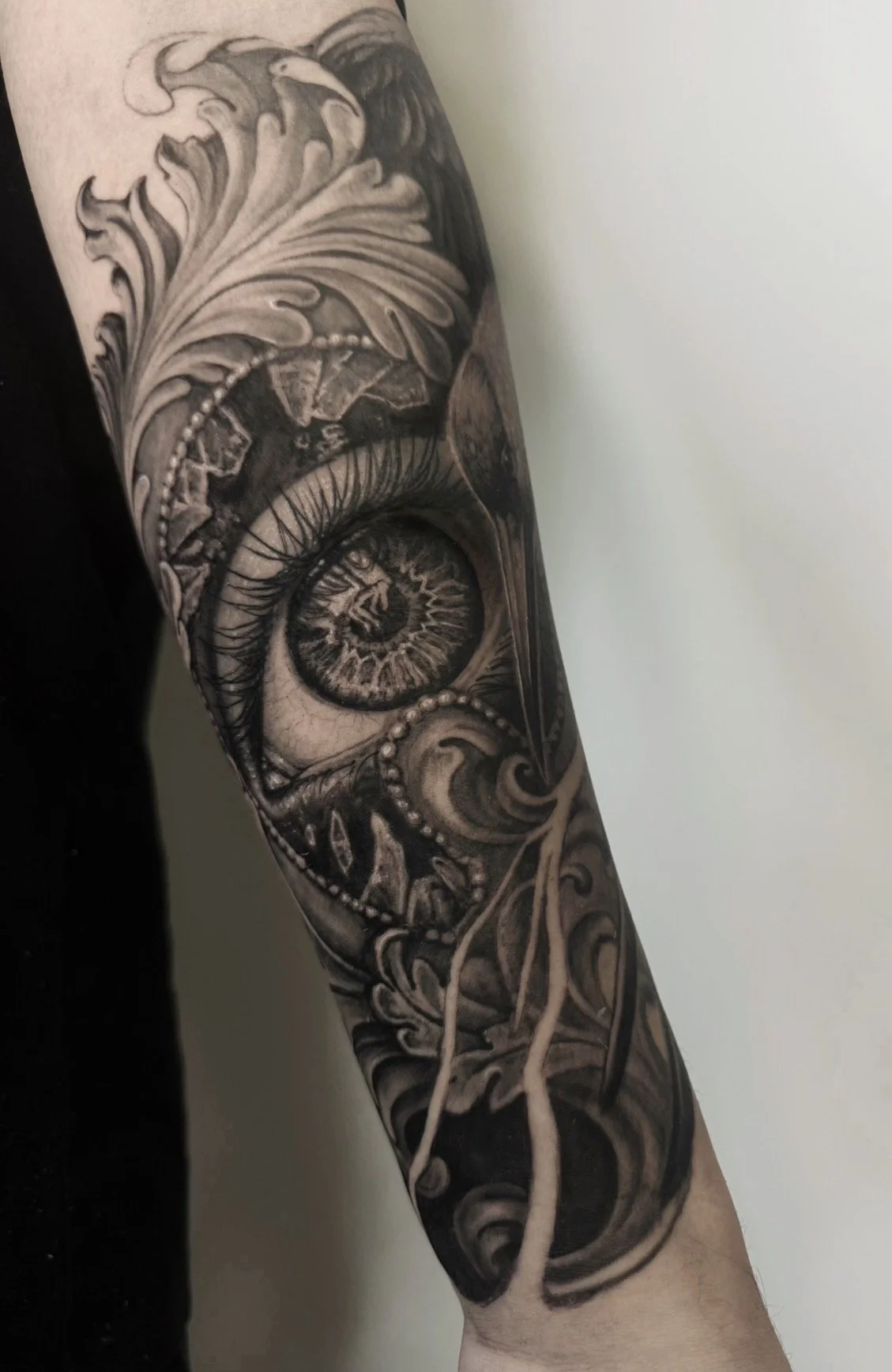 A black and gray tattoo on a person's arm featuring an eye, ornate swirling patterns, and a lion's head at the top.