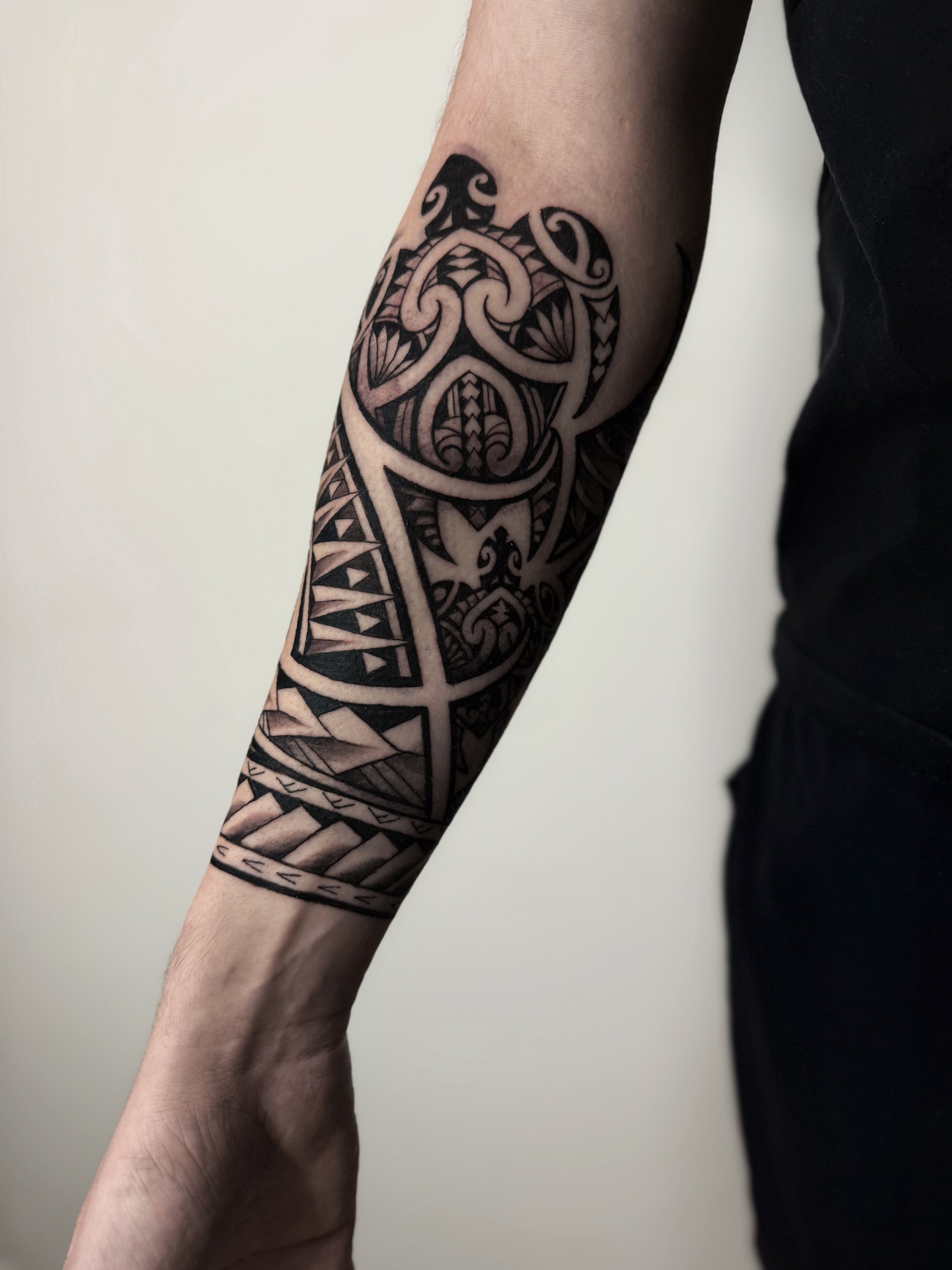 A person's forearm with a black and grey Polynesian-style tattoo featuring intricate tribal patterns and symbols.