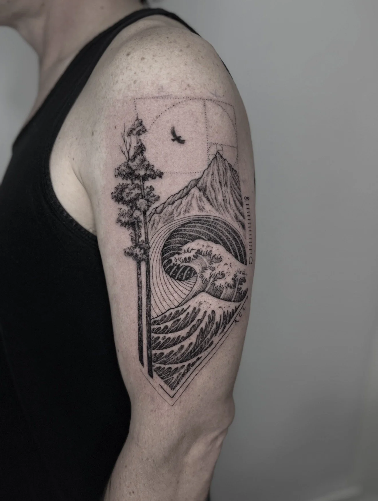 Tattoo of a mountain, a tree, a bird, and ocean waves, with geometric elements, on a person's upper arm.