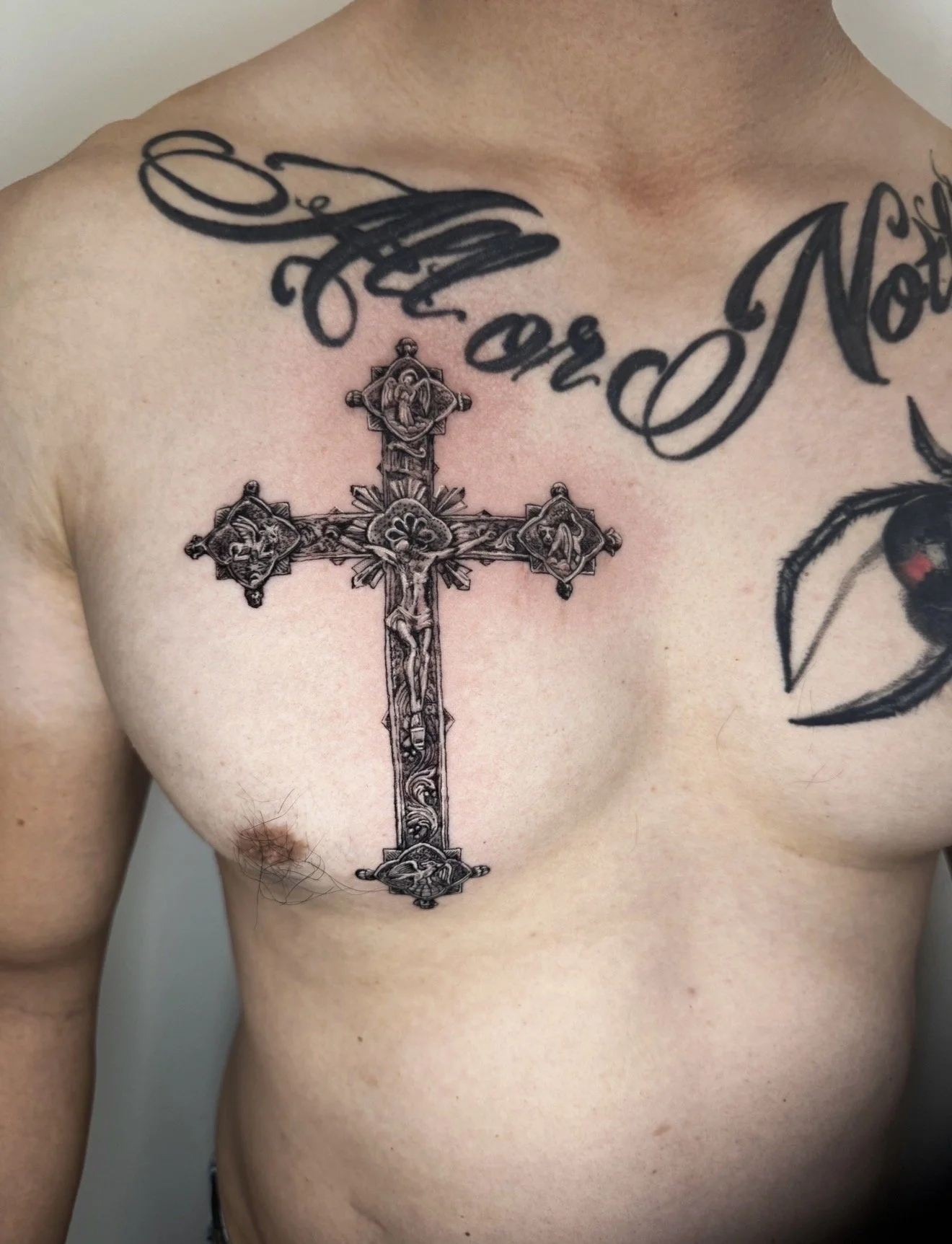 A tattoo of a detailed crucifix cross on a person's chest, with the person's skin visible, and part of a tattooed phrase reading 'Not' and other words visible.