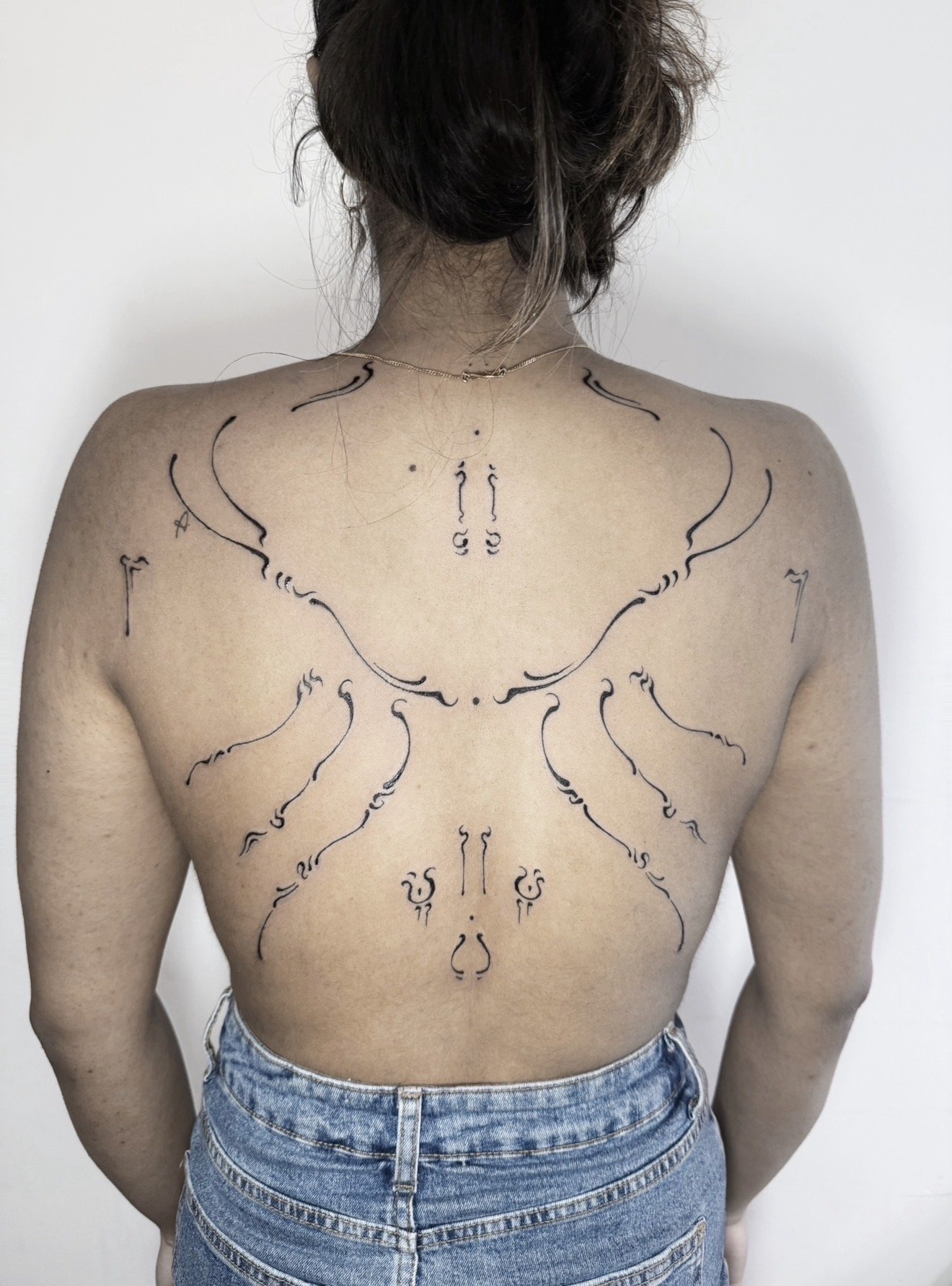 A woman with her back facing the camera has black tattoo outlines of spiritual symbols or runes all over her back and shoulders, and she is wearing a gold chain necklace and blue jeans.