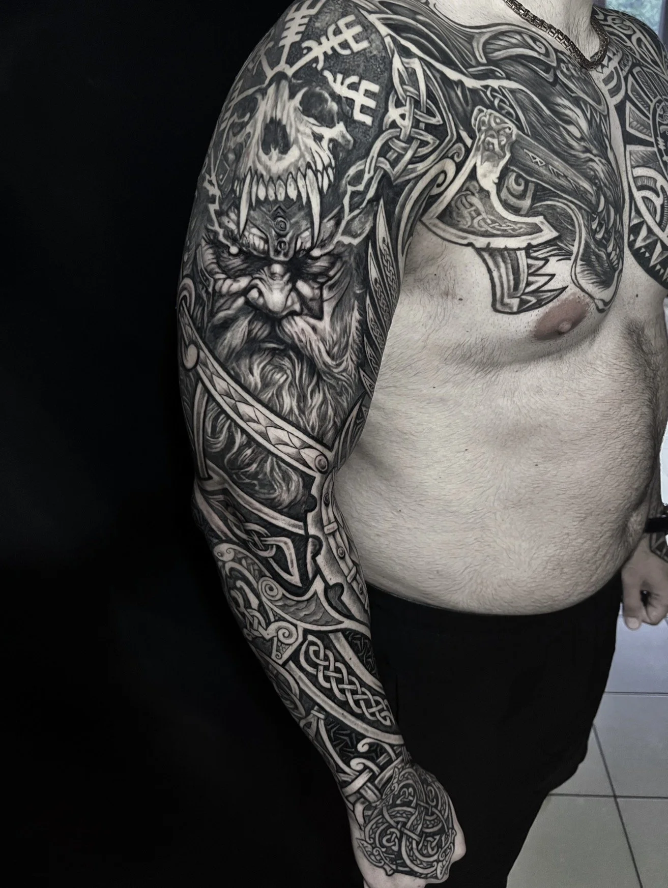 A person with extensive black and gray tattoos covering their left arm, shoulder, and chest. The tattoos include a Viking warrior with a beard and helmet, a fierce animal face, and various Celtic knot and tribal patterns.
