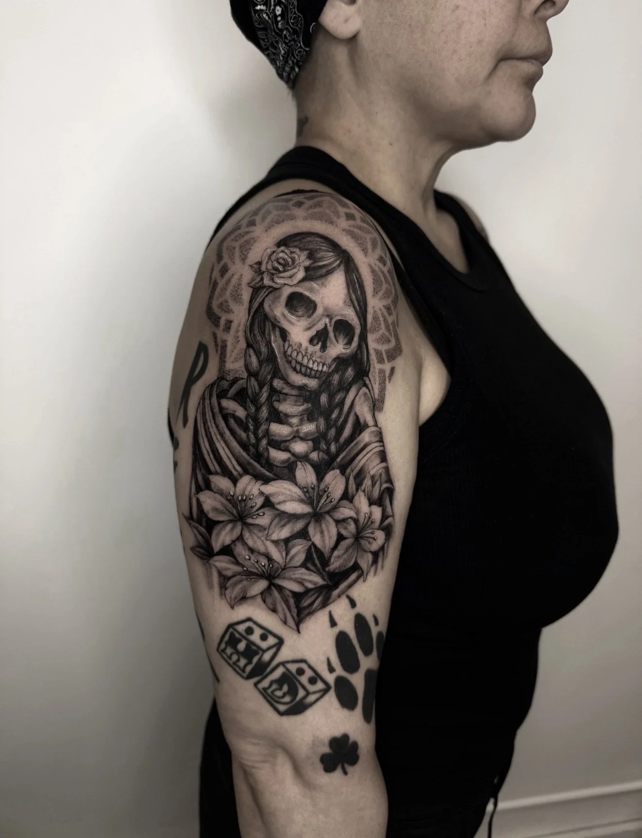 A woman with a black and white bandana on her head shows her right shoulder and upper arm, which has a black and gray tattoo of a skeleton woman with braided hair, flowers, and decorative elements.