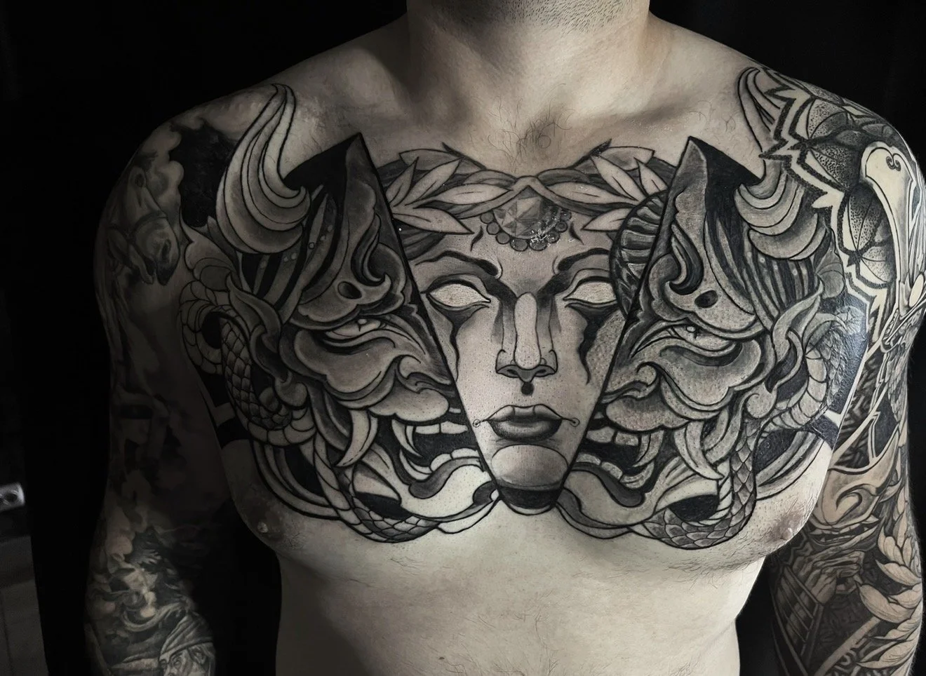 Black and gray chest tattoo of a woman's face with long flowing hair, surrounded by fierce animal faces such as a lion and a snake, with intricate details.
