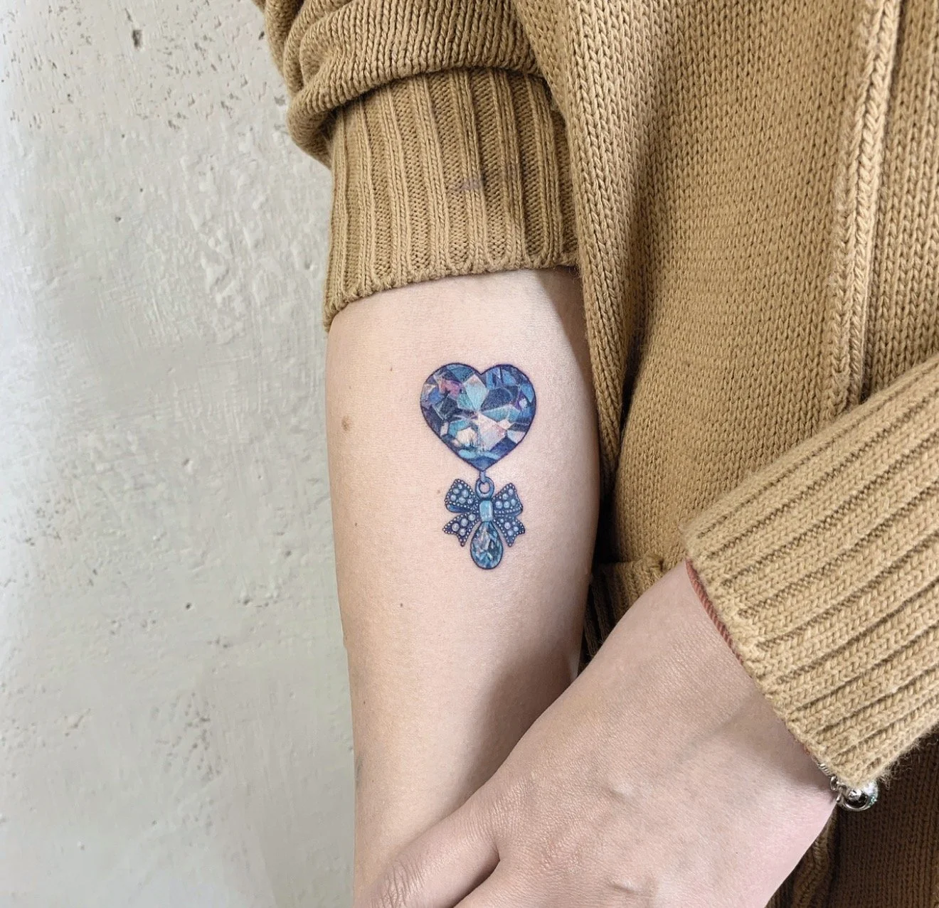 A tattoo of a blue and purple heart-shaped gemstone with a dangling bow-shaped charm on a person's upper arm, who is wearing a tan sweater.